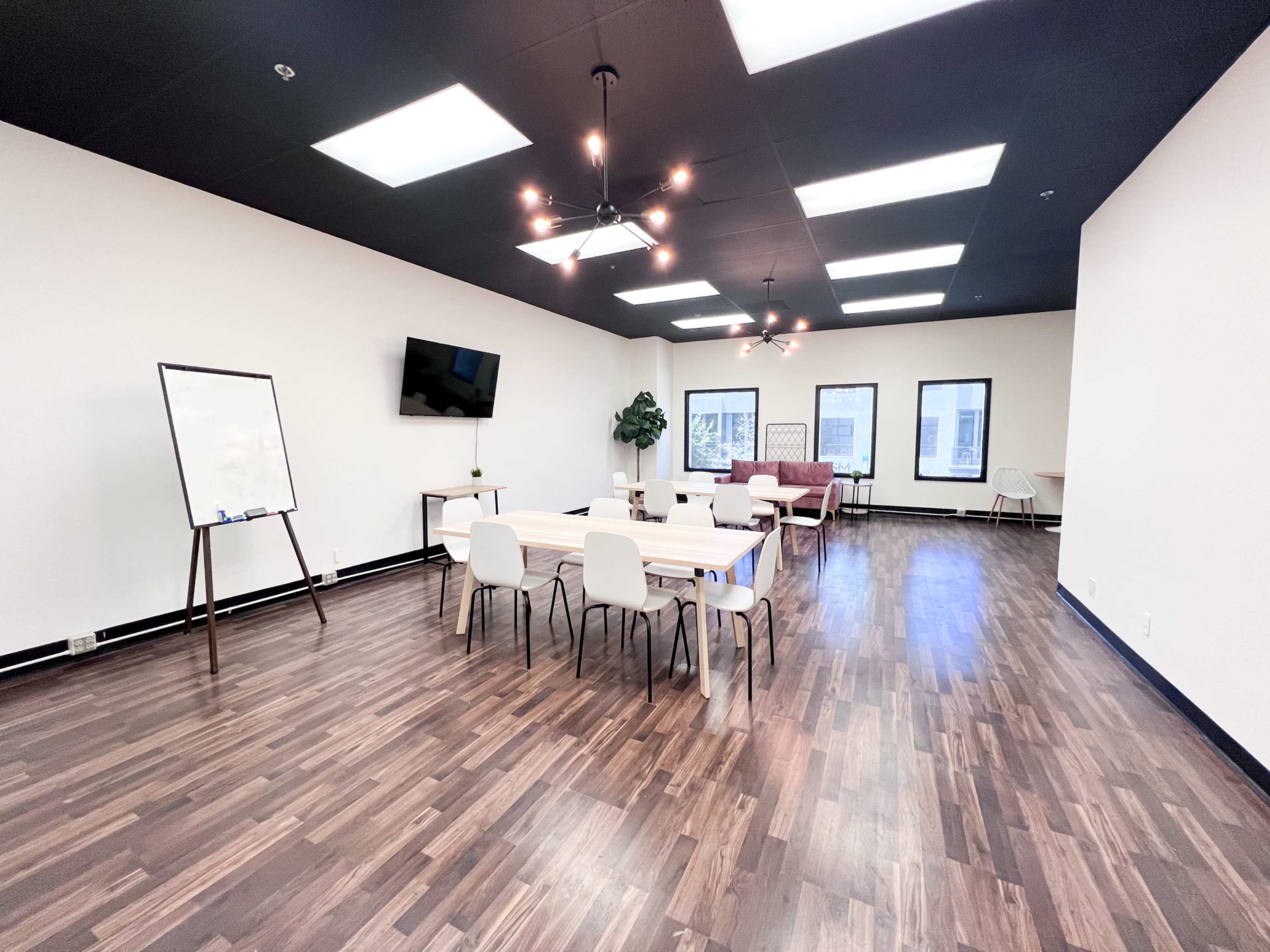 A spacious meeting room with wooden floors, several tables and chairs, a whiteboard, and a television mounted on the wall.