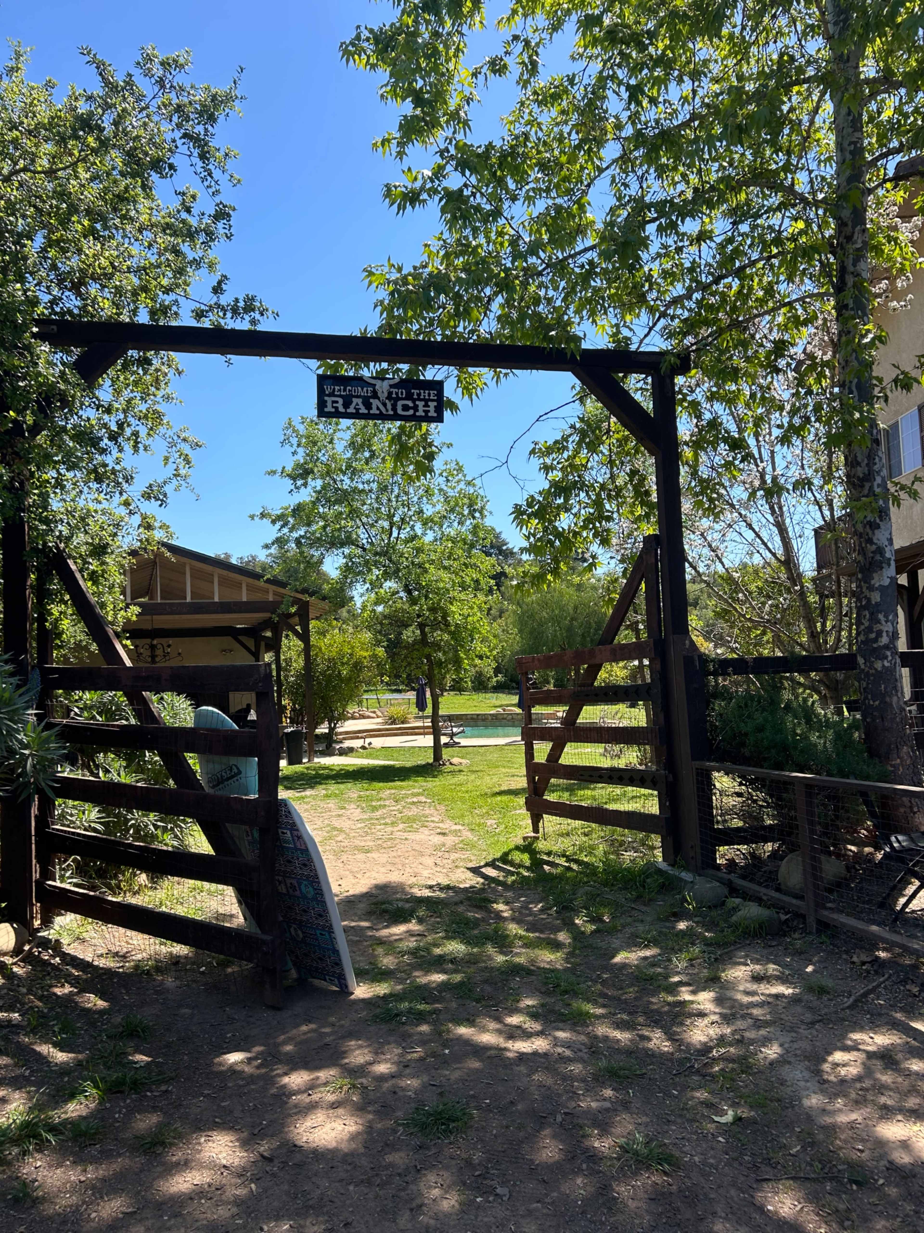 A wooden entrance gate with a sign that reads "Ranch" leads to a grassy area surrounded by trees and structures.