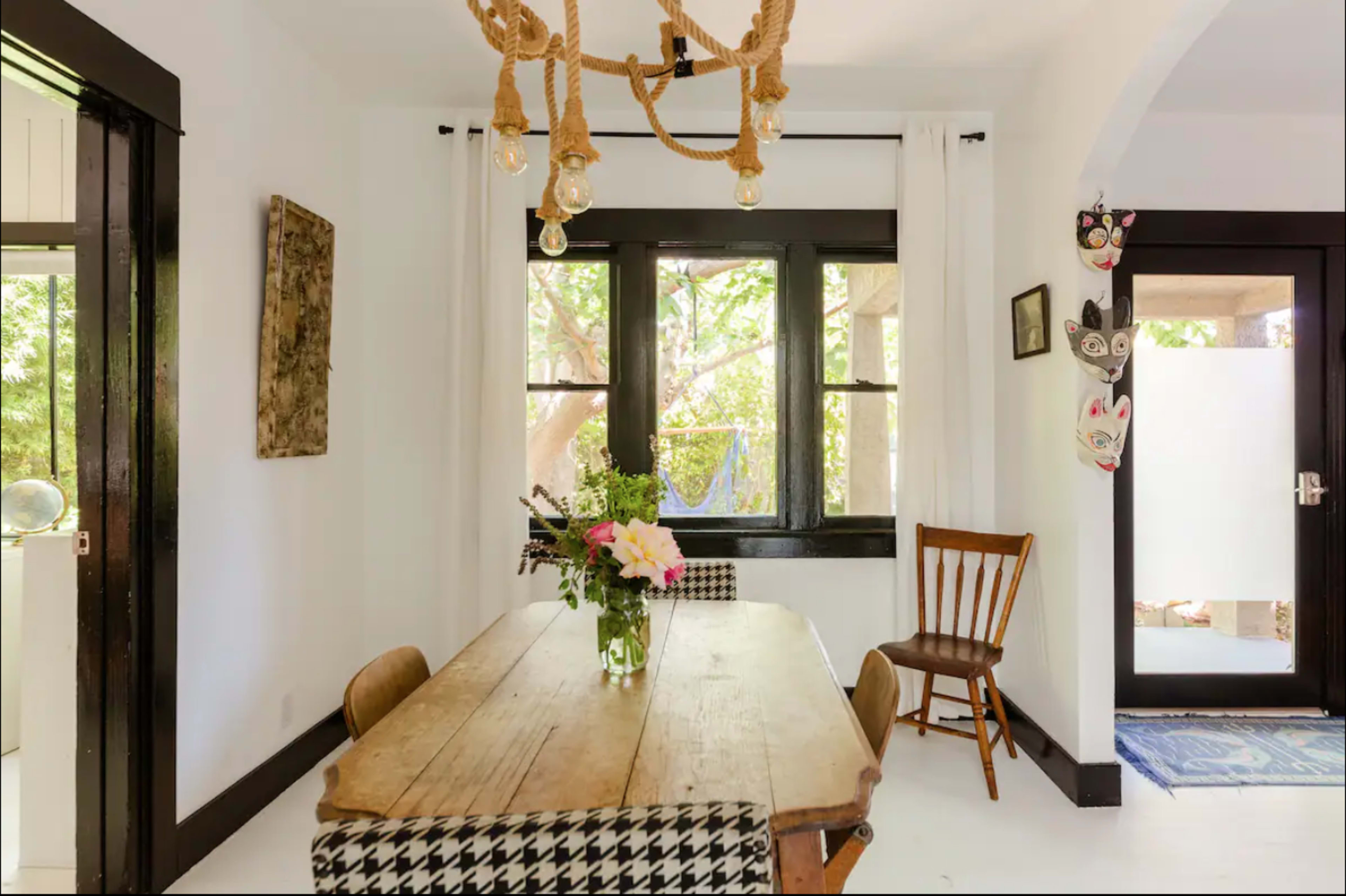 The image features a minimalist dining area with a wooden table, a flower arrangement in the center, and large windows allowing natural light to enter.