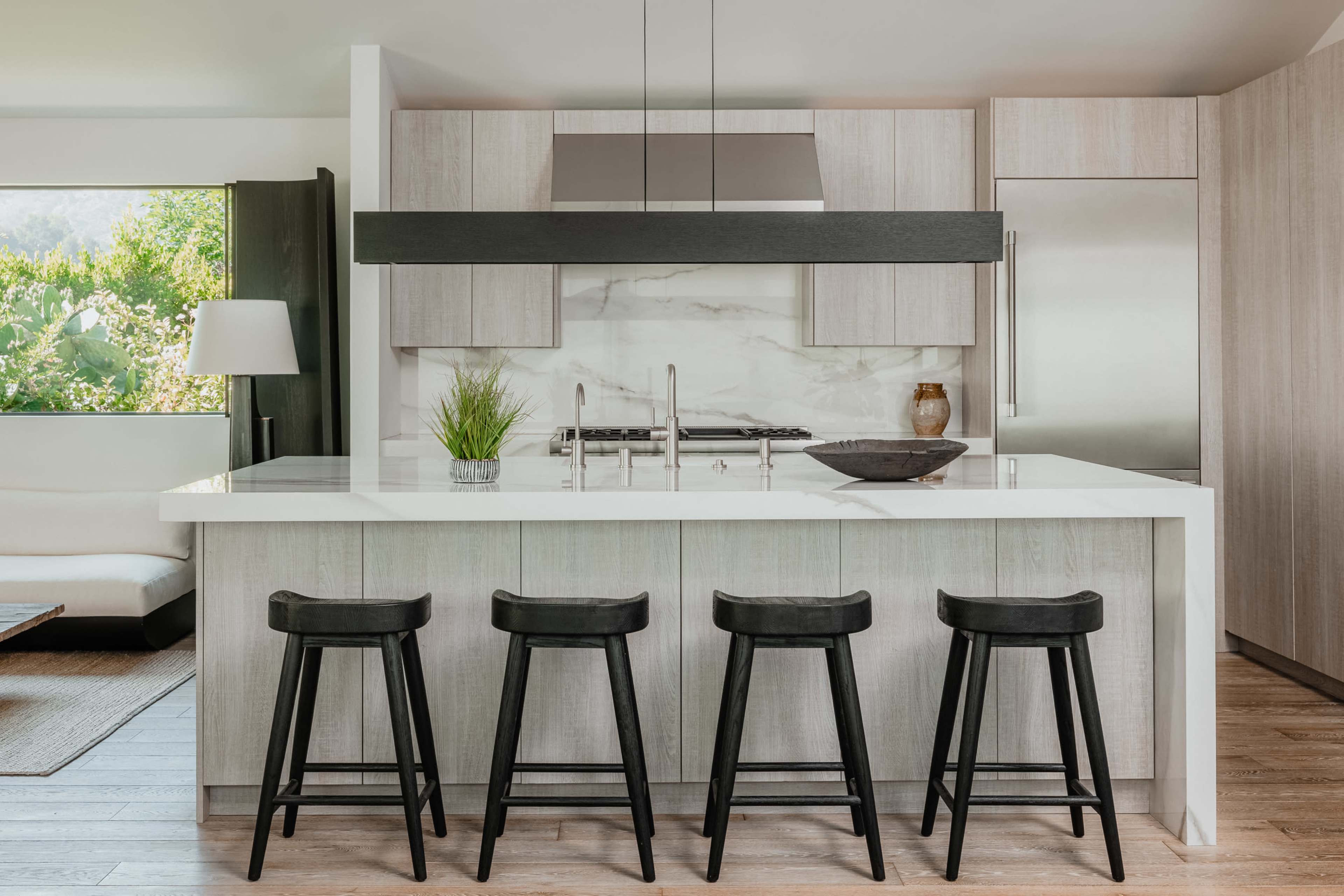 The image shows a modern kitchen with a central island featuring four black stools, a stainless steel refrigerator, and a marble backsplash.