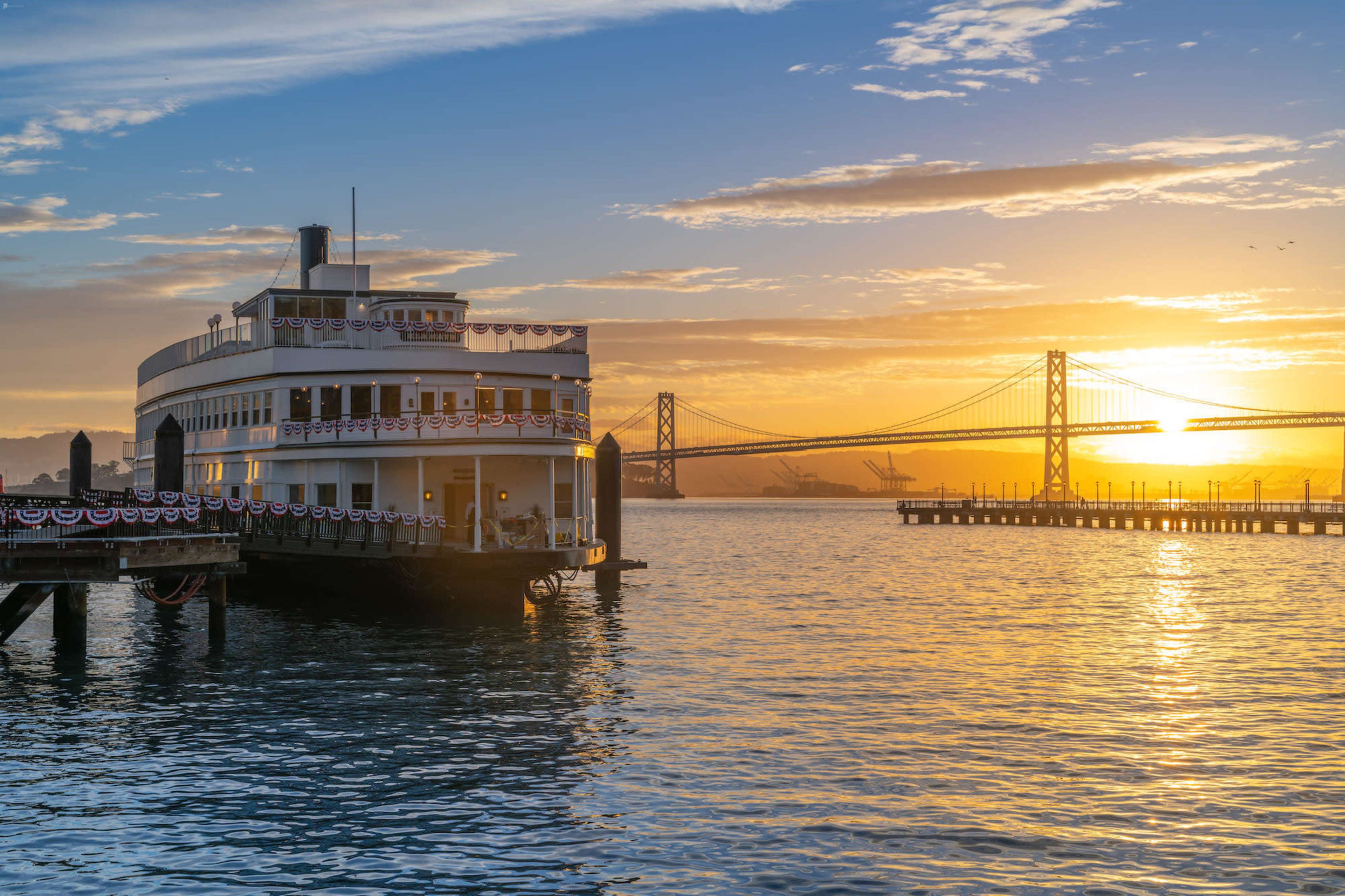 A large boat adorned with decorations is docked at a pier during sunset, with the Bay Bridge visible in the background.