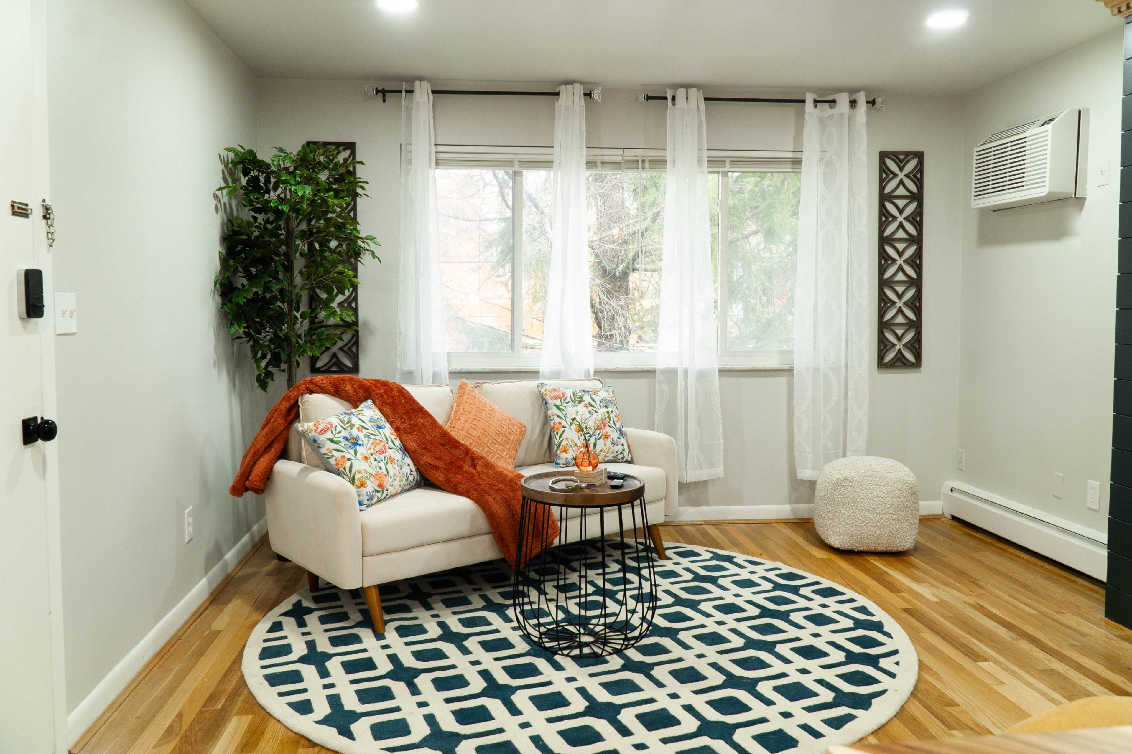 A living room with a light-colored couch, decorative throw pillows, a round rug, a coffee table, and a large window with sheer curtains.