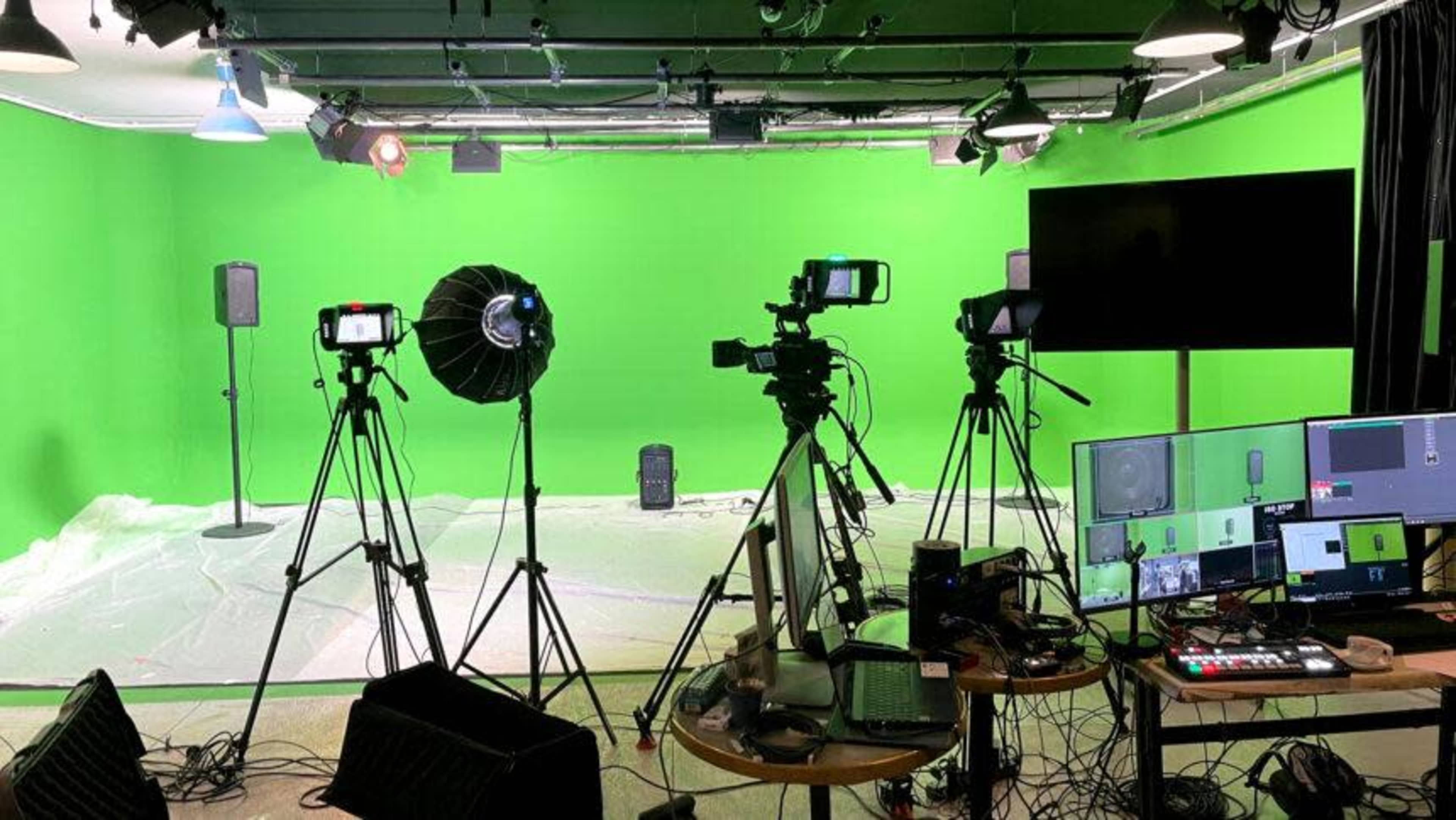 The image shows a green screen studio equipped with multiple cameras, lighting equipment, and a control panel for video production.