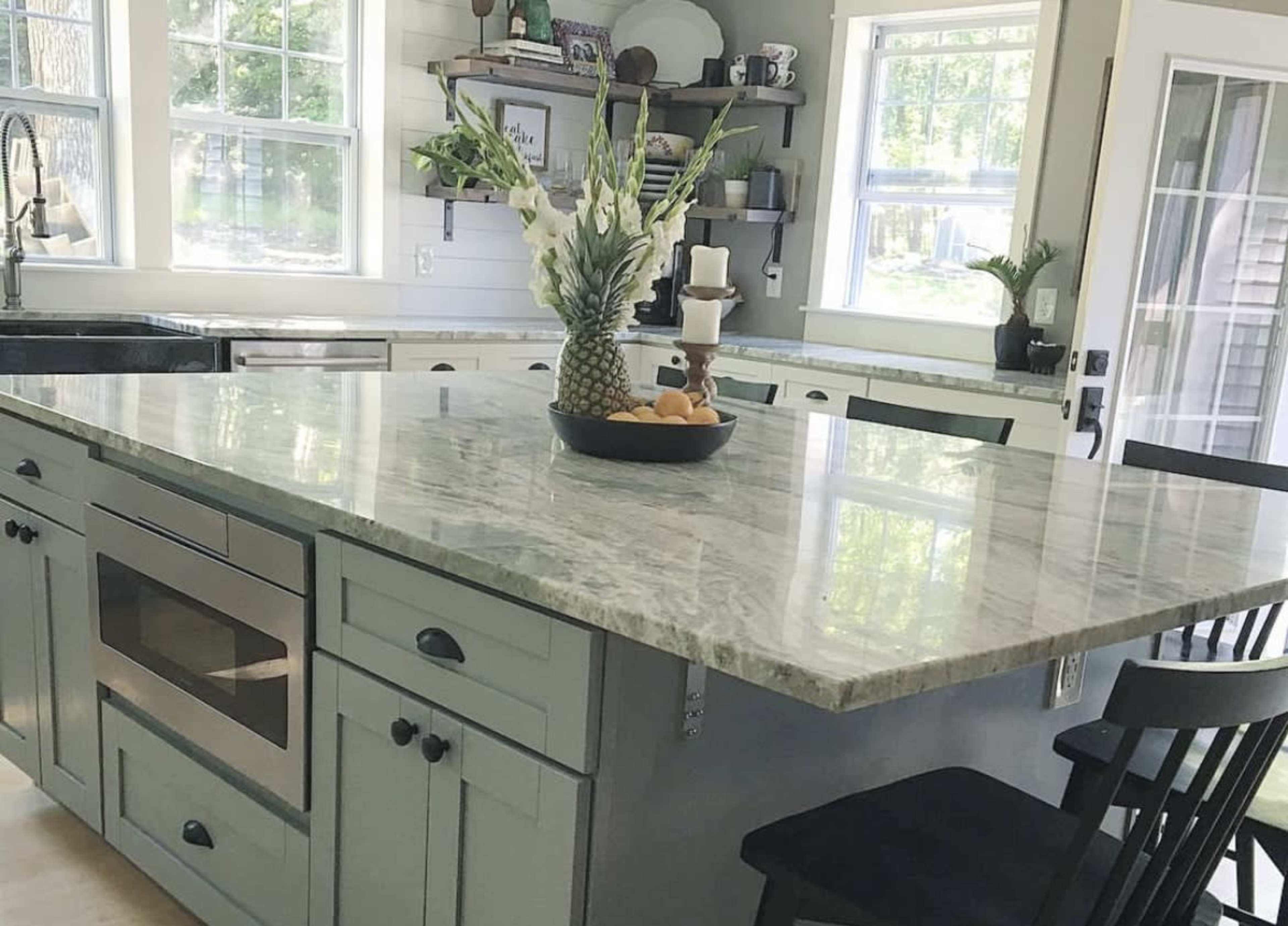 A modern kitchen features a large island with a speckled countertop, surrounded by black chairs and filled with a bowl of fruit.
