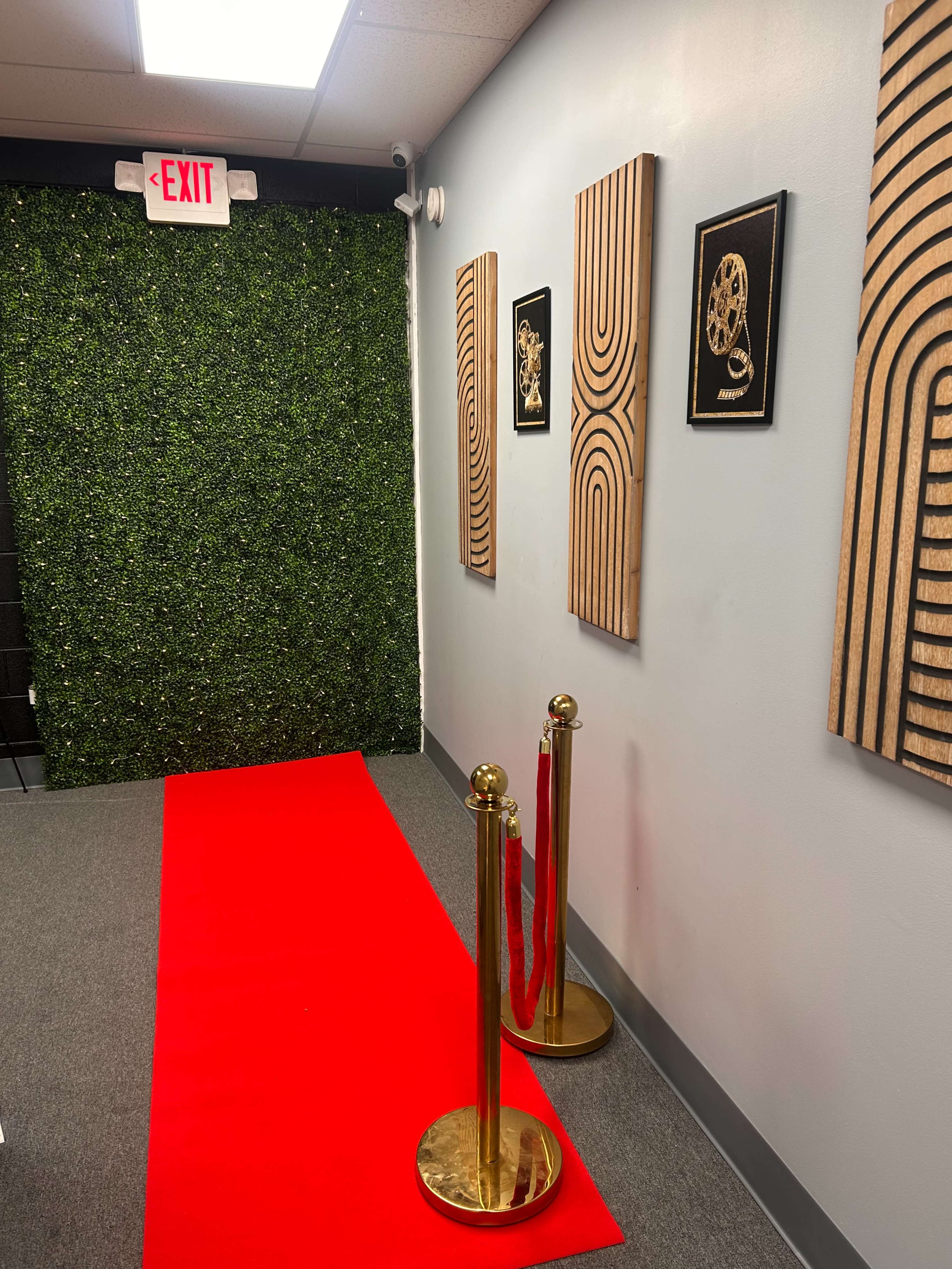 The image shows a hallway with a red carpet, gold stanchions with red ropes, and wooden wall art, leading to a green, leafy wall and an exit sign.