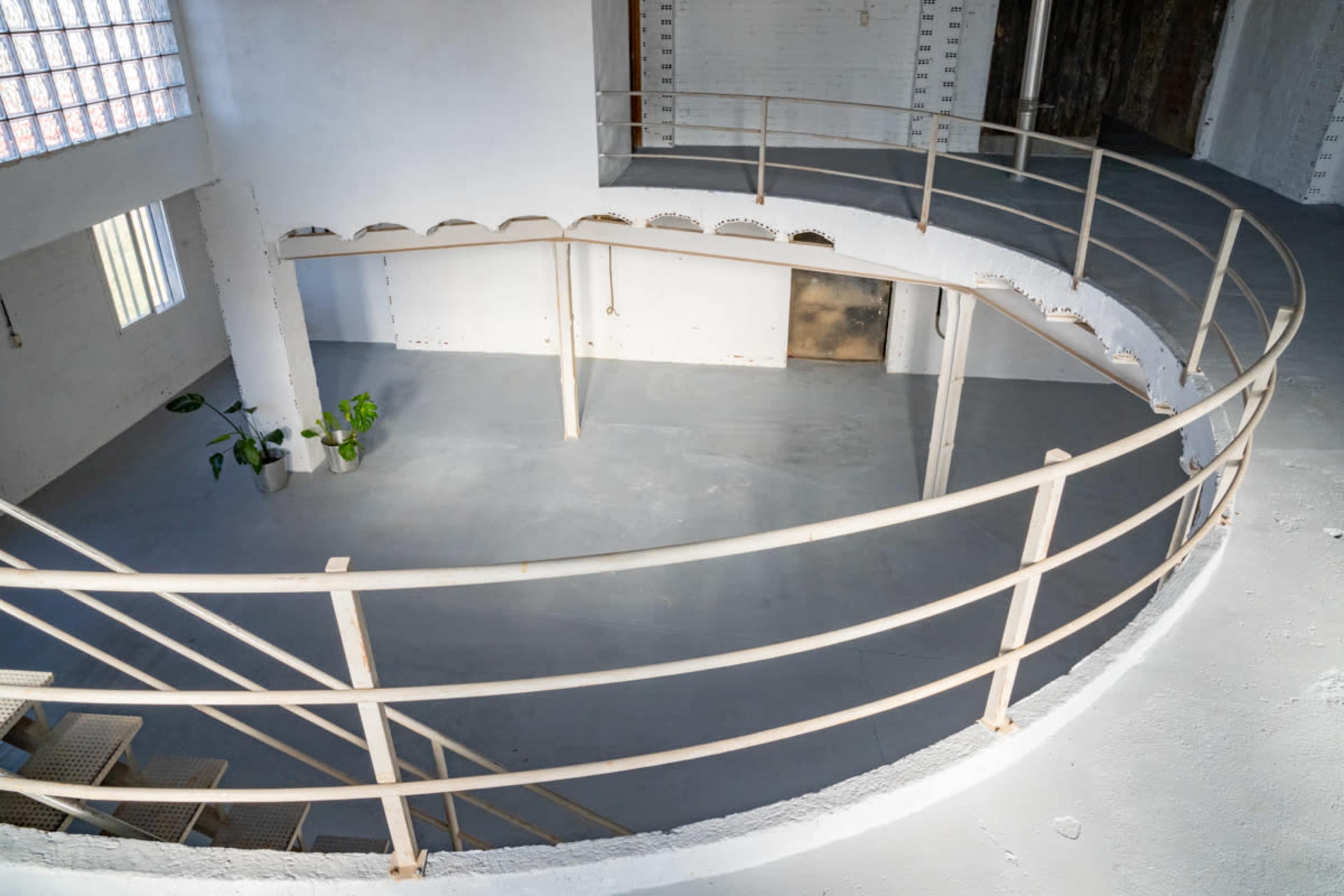The image shows a curved metal railing surrounding a spacious, empty interior with concrete walls and a gray floor.