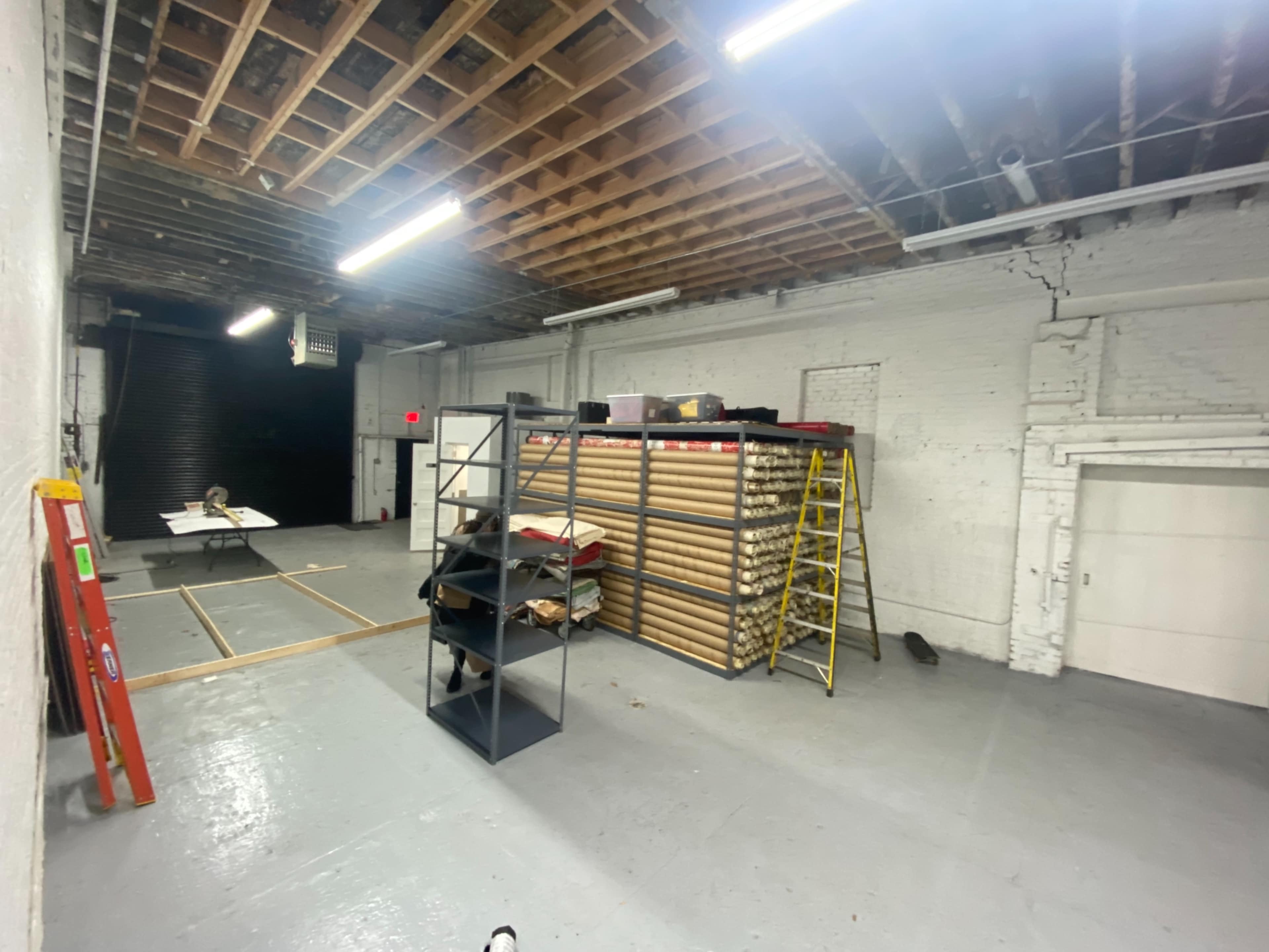 A spacious, unfinished room with exposed beams, a shelving unit stacked with materials, a ladder, and a construction setup in the corner.