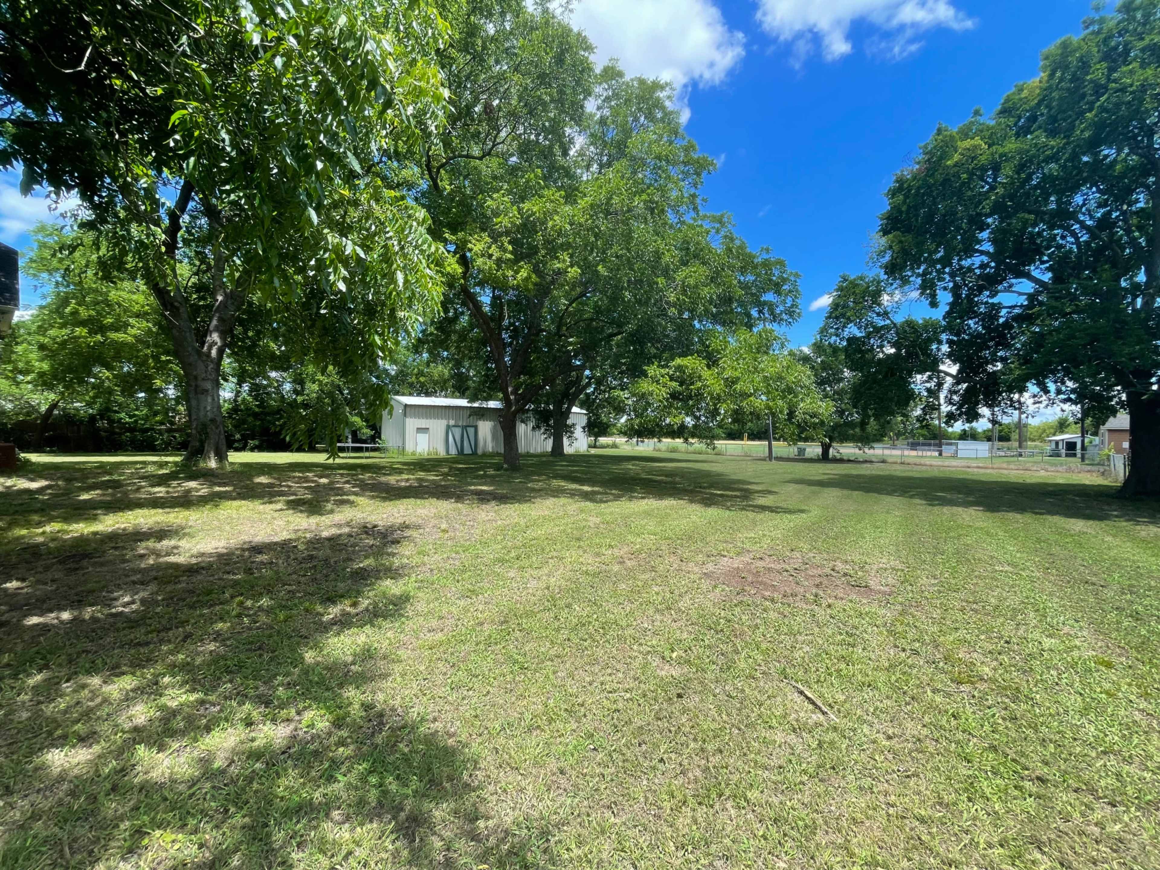 The image shows a spacious grassy area with several trees and a white building in the background under a clear blue sky.