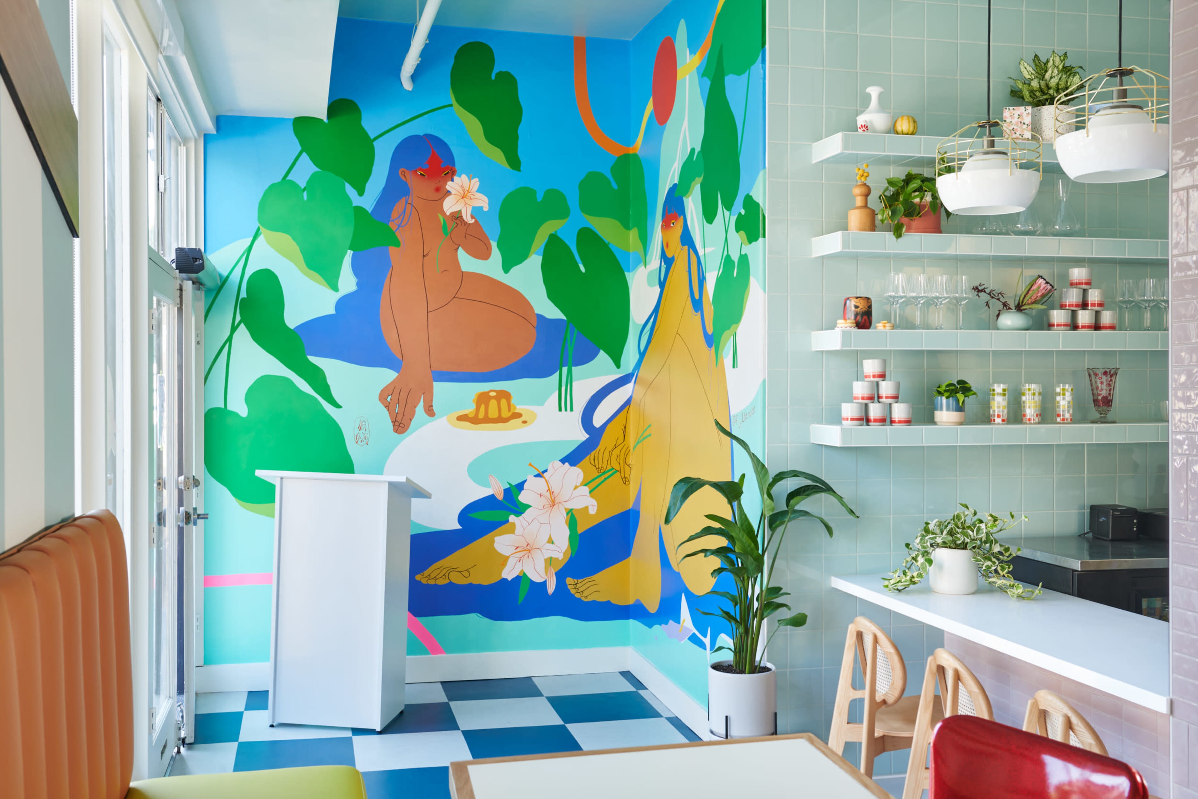 A brightly colored mural featuring stylized figures surrounded by tropical foliage on a wall in a modern cafe, with a clean, minimalist interior design.