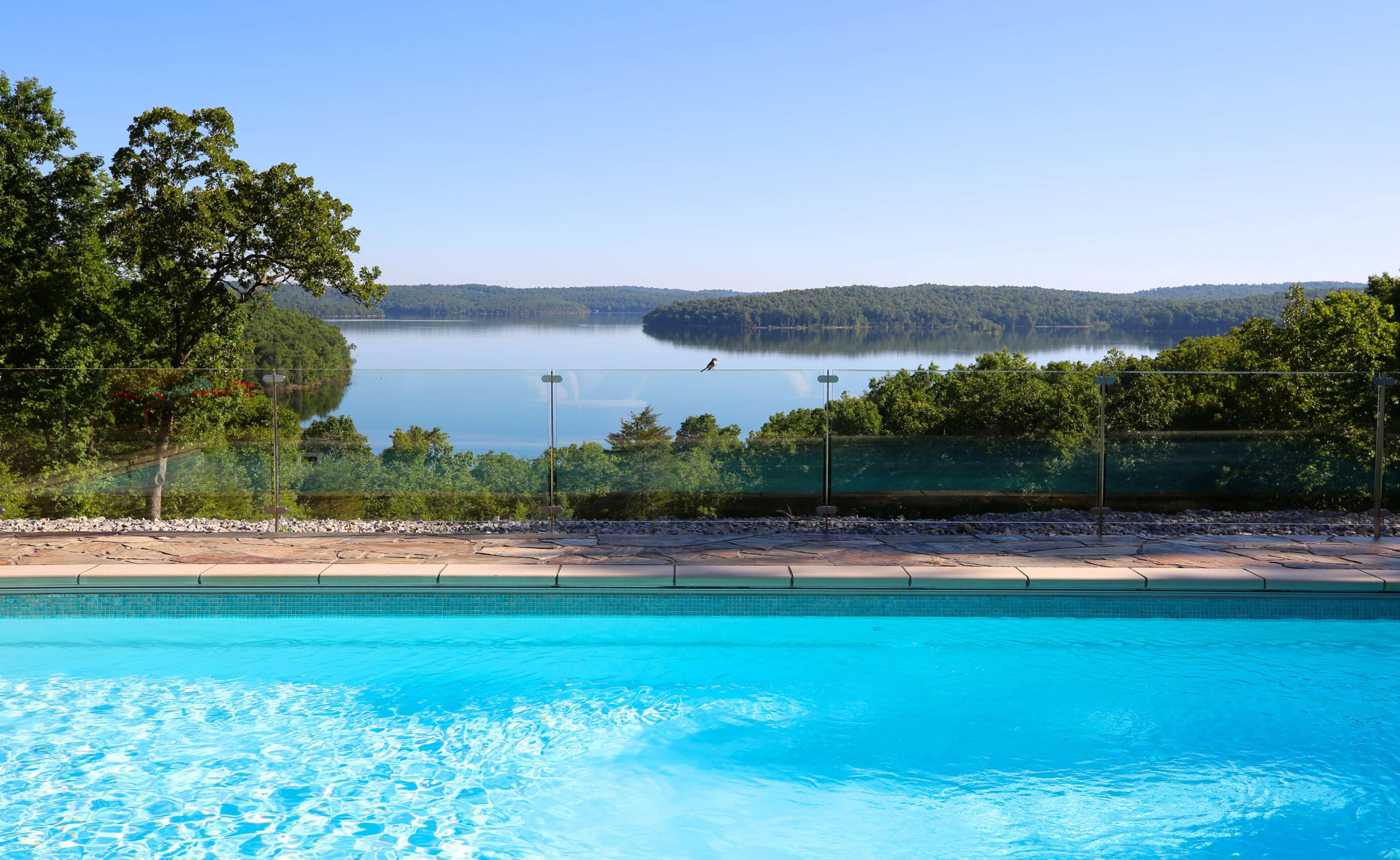 The image shows a swimming pool with clear blue water overlooking a calm lake surrounded by greenery and hills in the background.