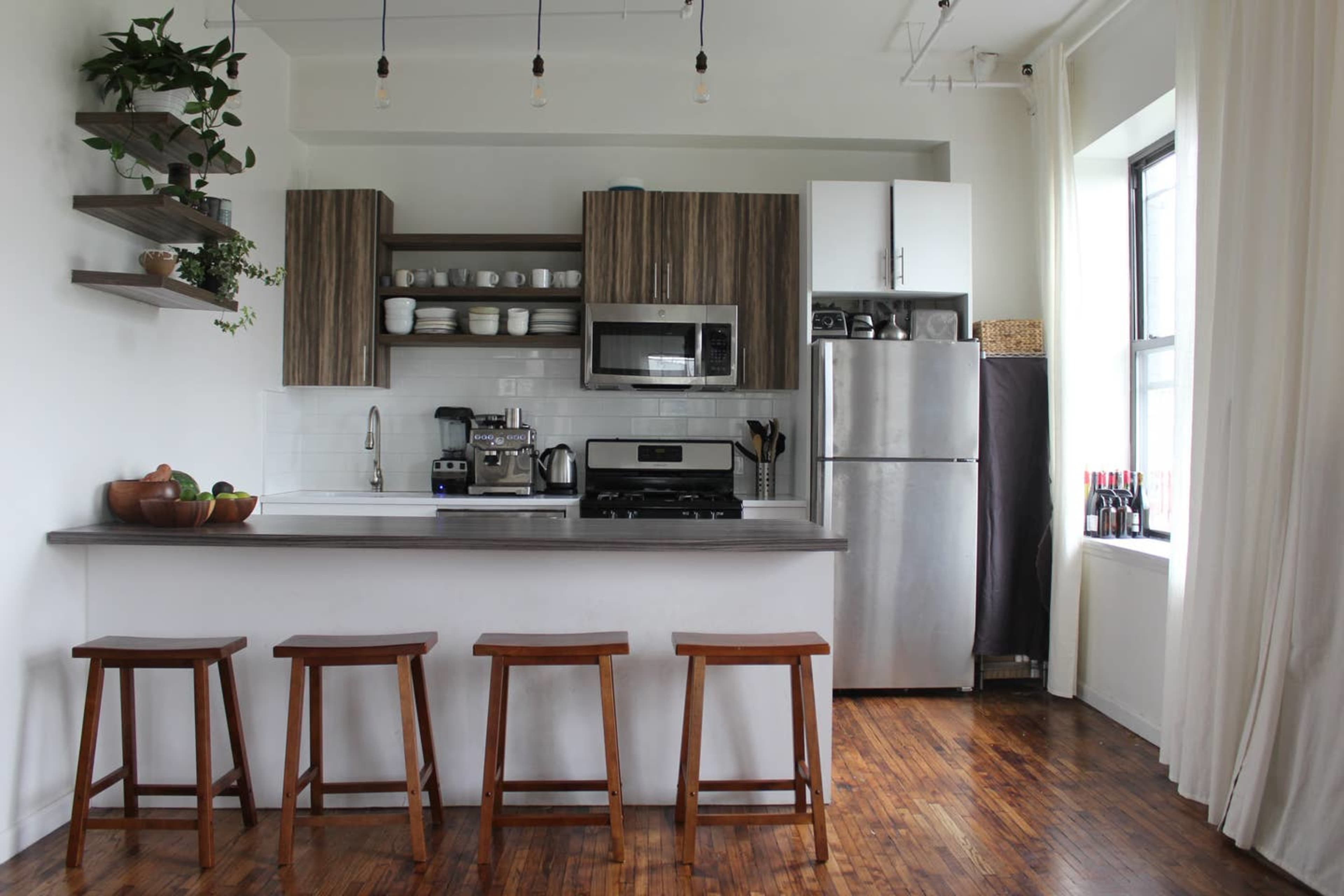 The kitchen features wooden shelves, modern appliances, and three stools lined up at a counter.