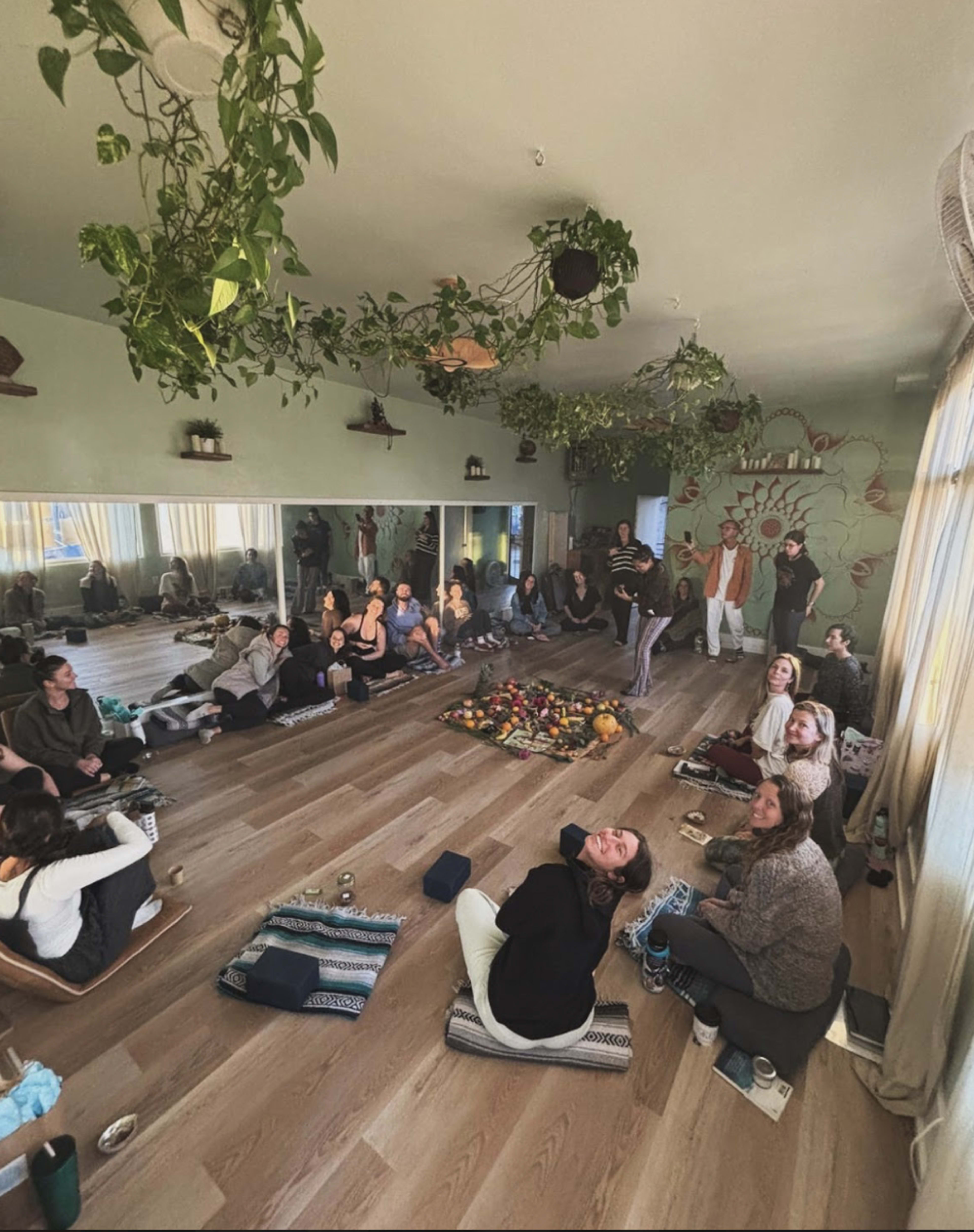 A group of people is seated on the floor of a brightly lit room filled with plants, participating in a workshop or gathering centered around a circular arrangement of fruits and candles.