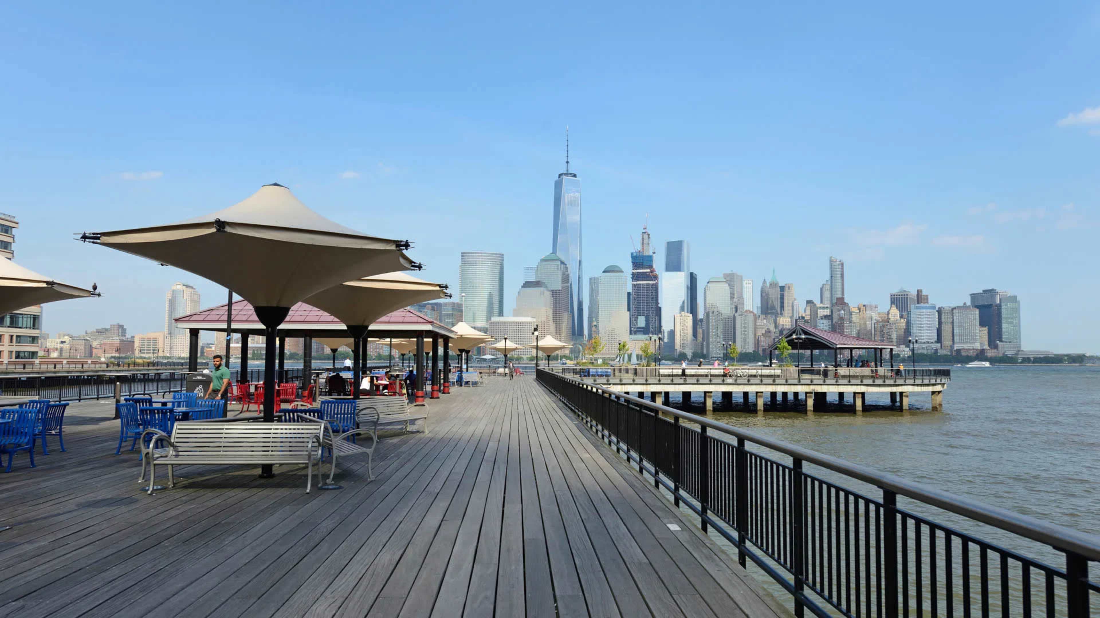 The image shows a wooden pier with shaded seating areas and a view of the Manhattan skyline, including One World Trade Center, in the background.