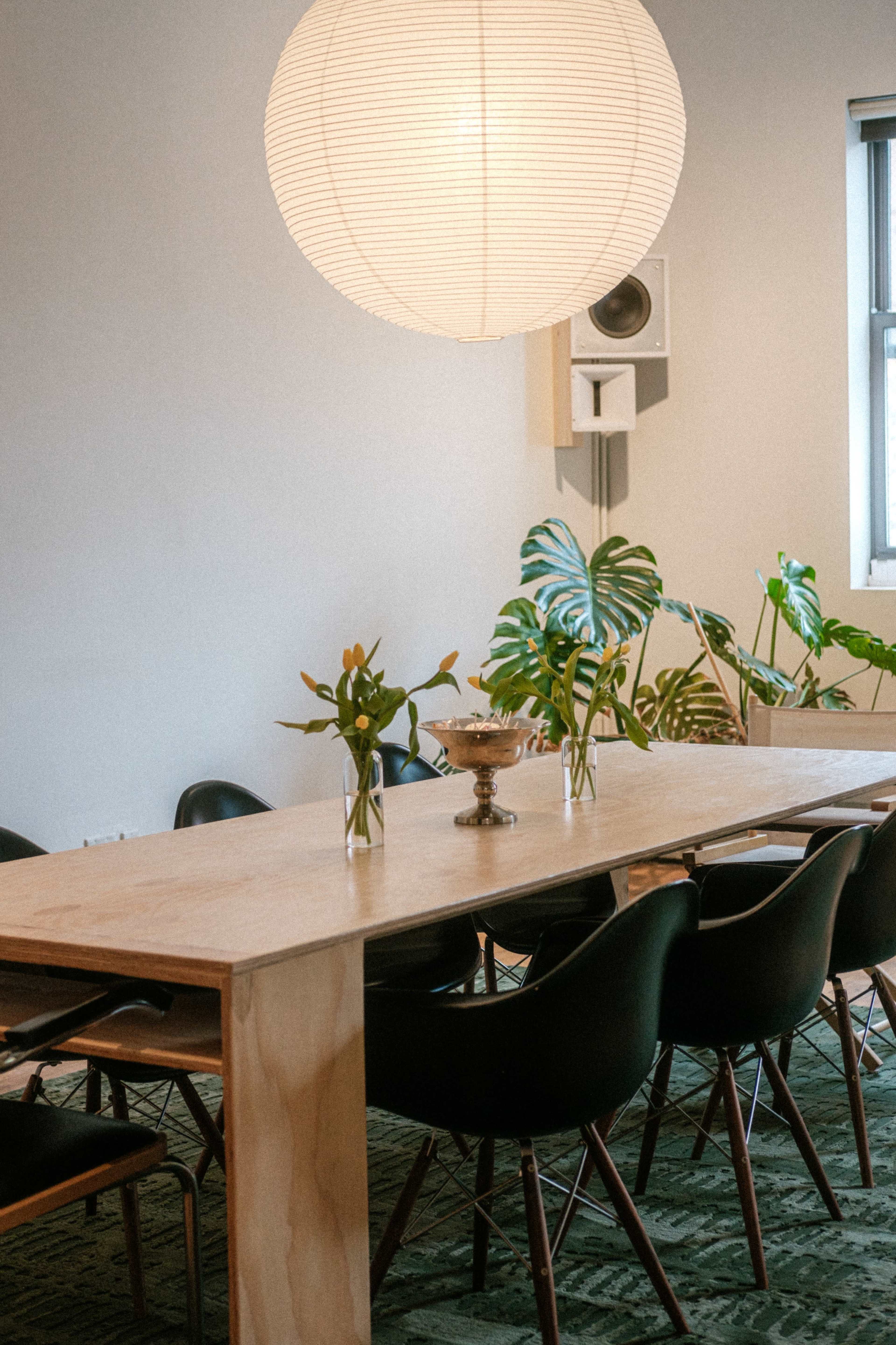 A long wooden dining table is set with vases of flowers and surrounded by black chairs in a room with a large pendant light and indoor plants.