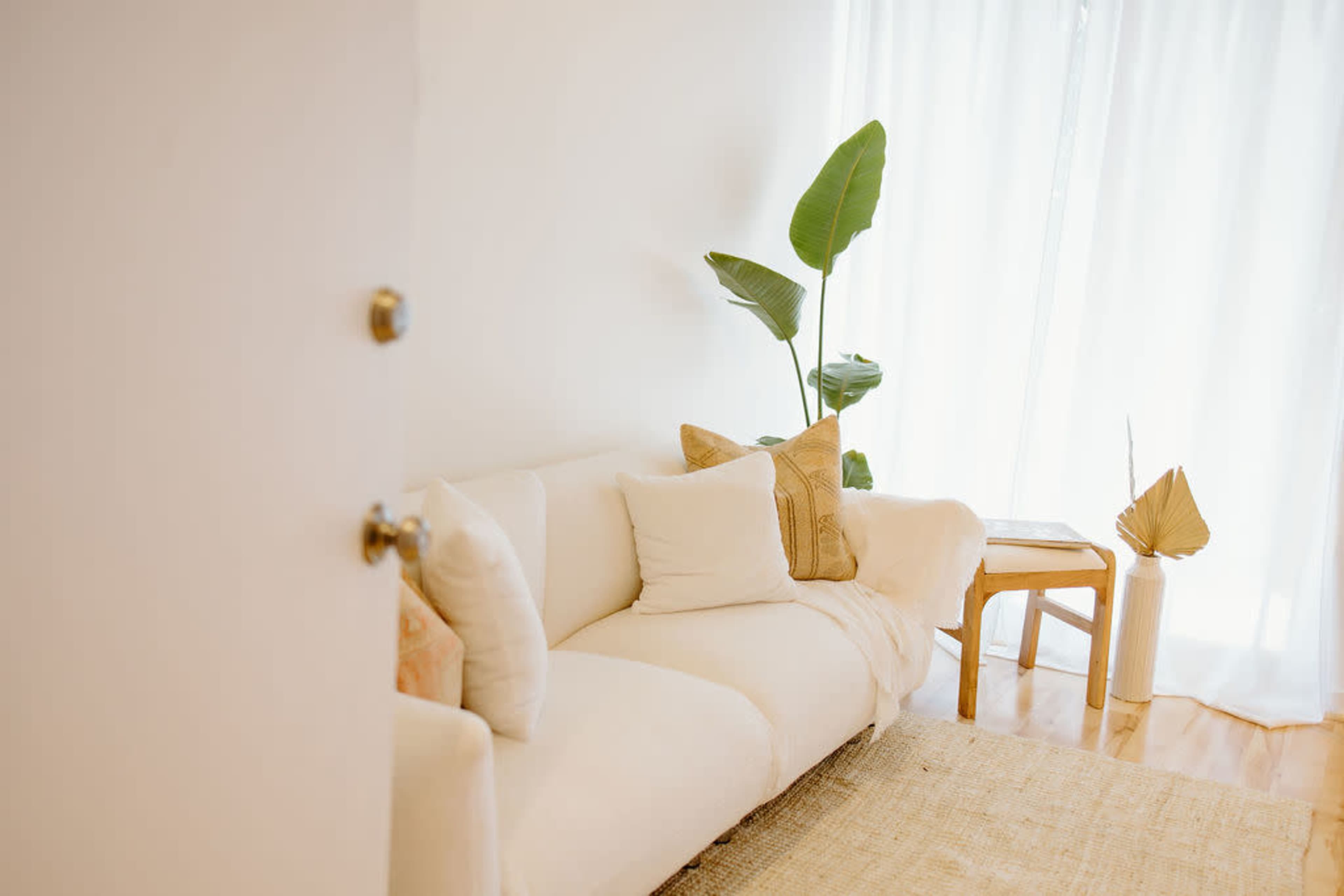 A white couch with decorative pillows is set beside a small side table near a window covered by sheer curtains, with a plant positioned nearby.