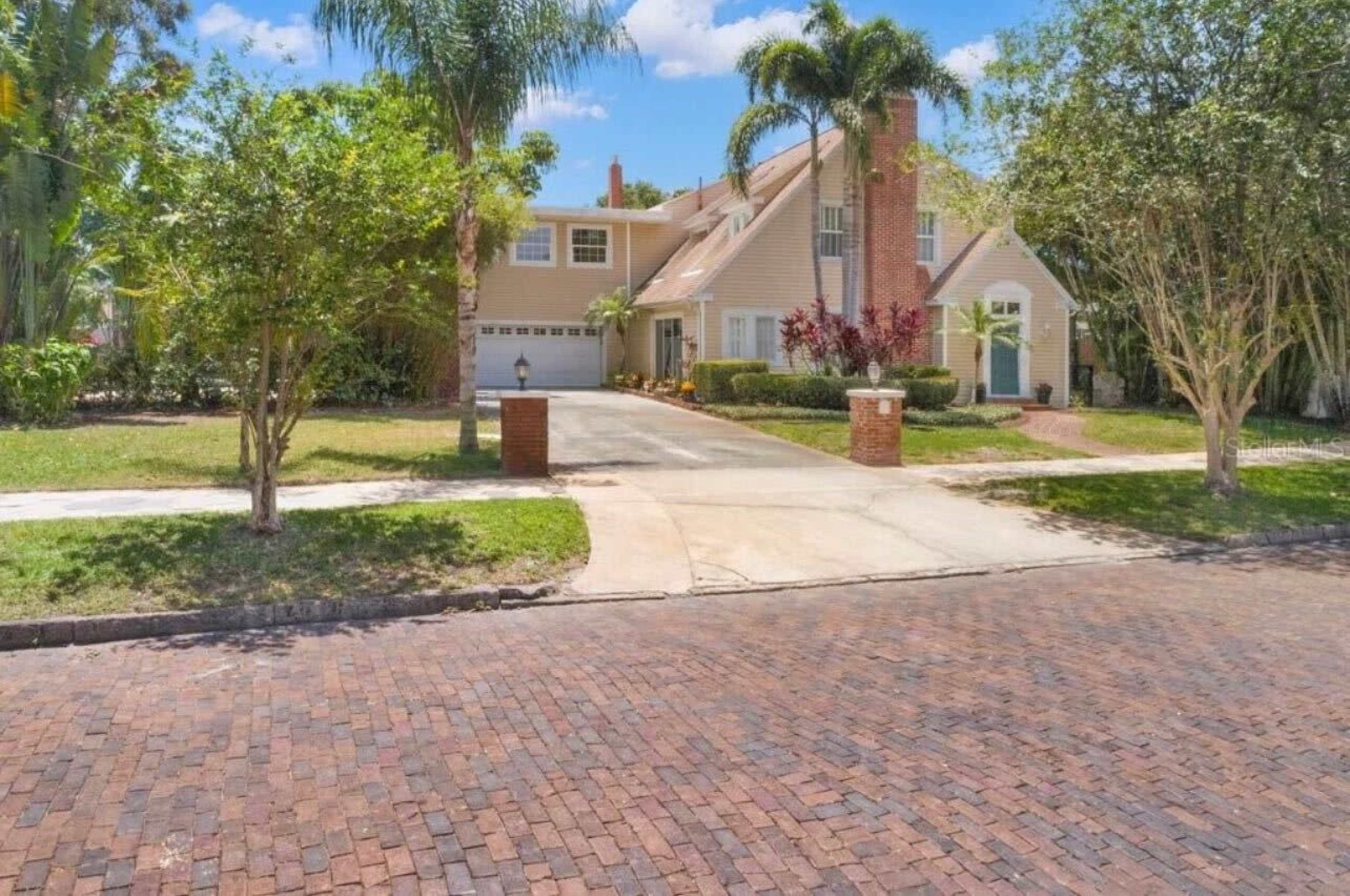 A two-story house with a brick driveway and landscaped yard is situated on a quiet street.