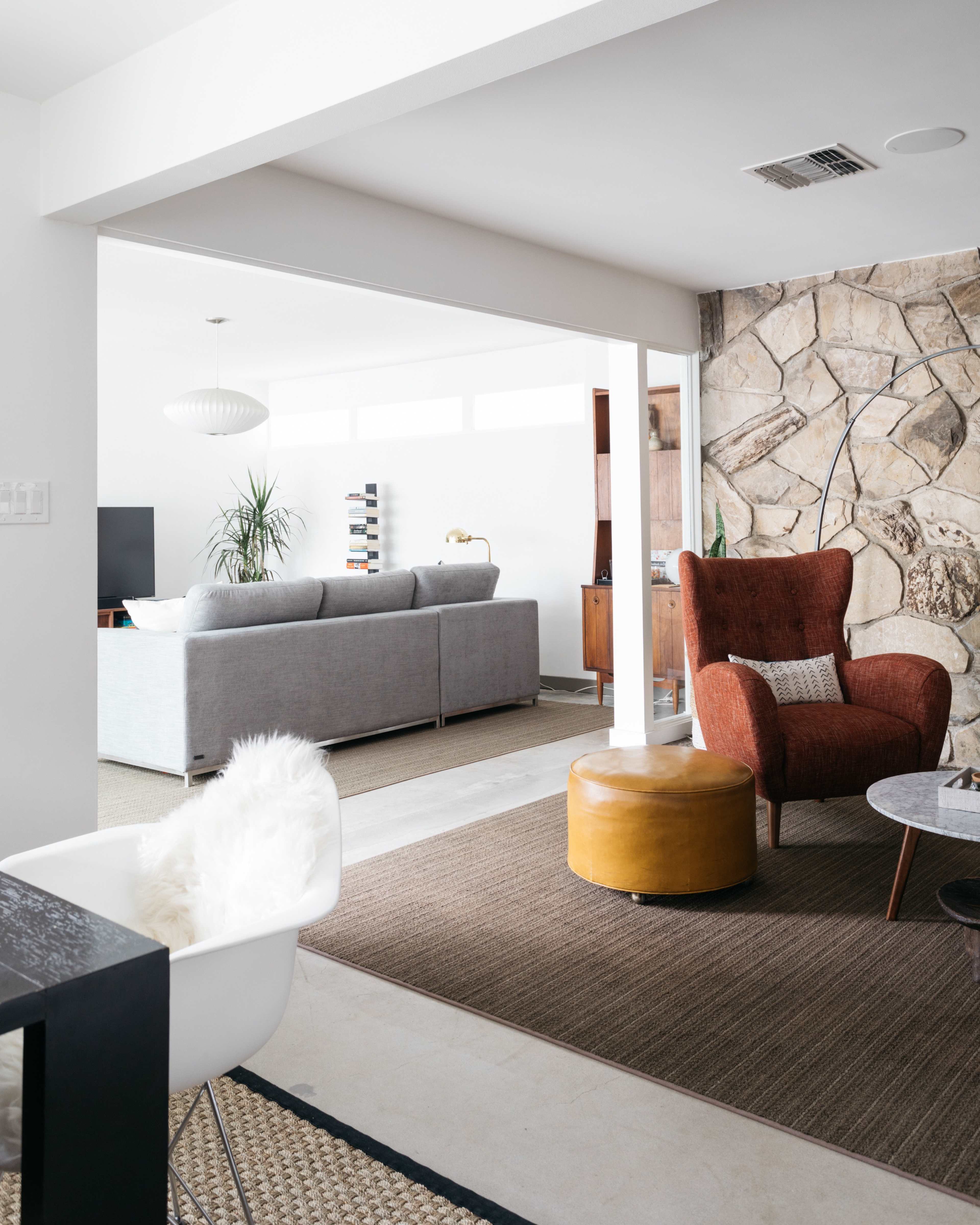 A modern, open-concept living room with a gray sofa, a brown accent chair, and a stone feature wall, leading to a well-lit space with plants and furniture.