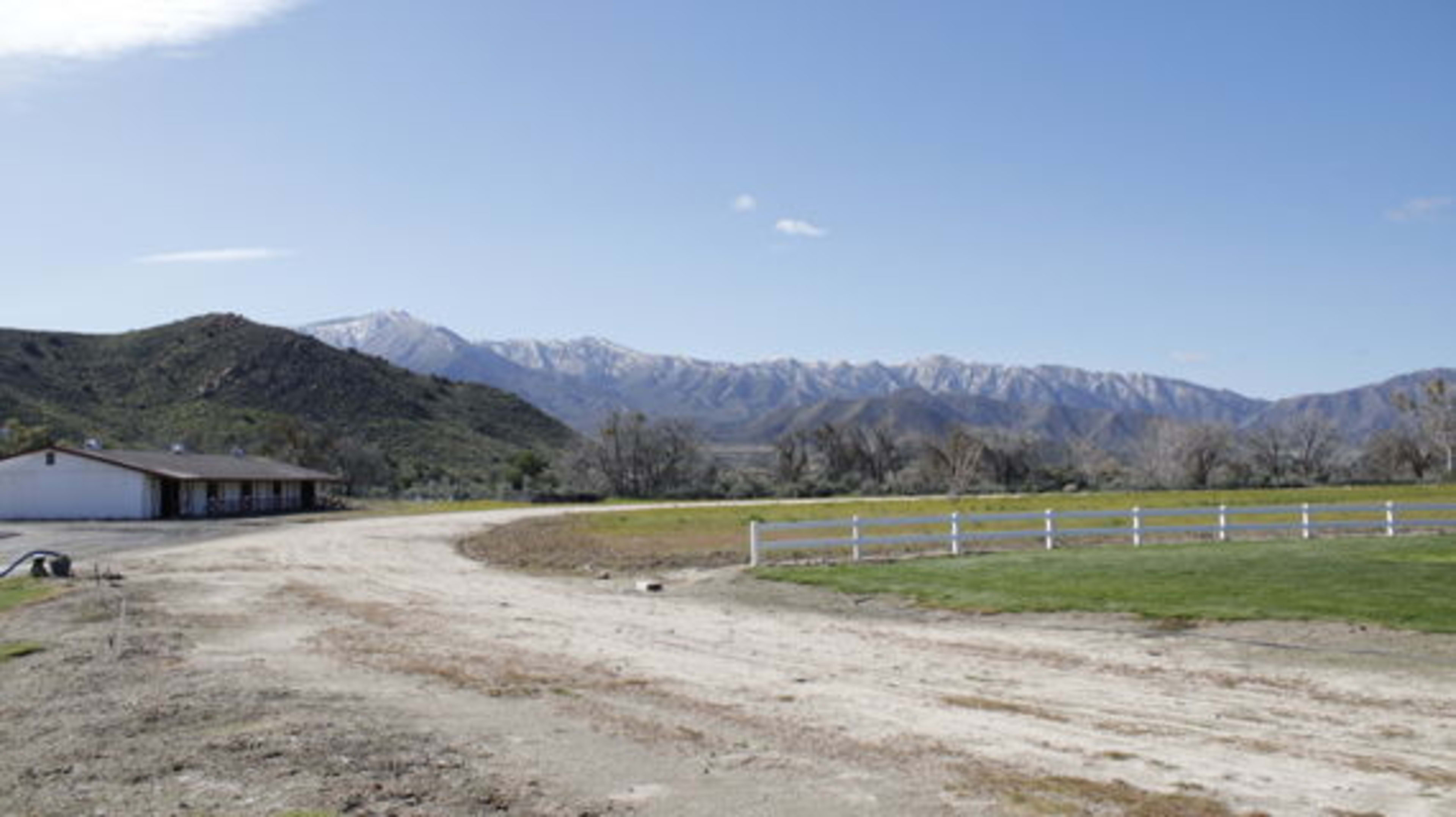 A gravel road leads to a ranch house with mountains in the background, some of which are capped with snow.