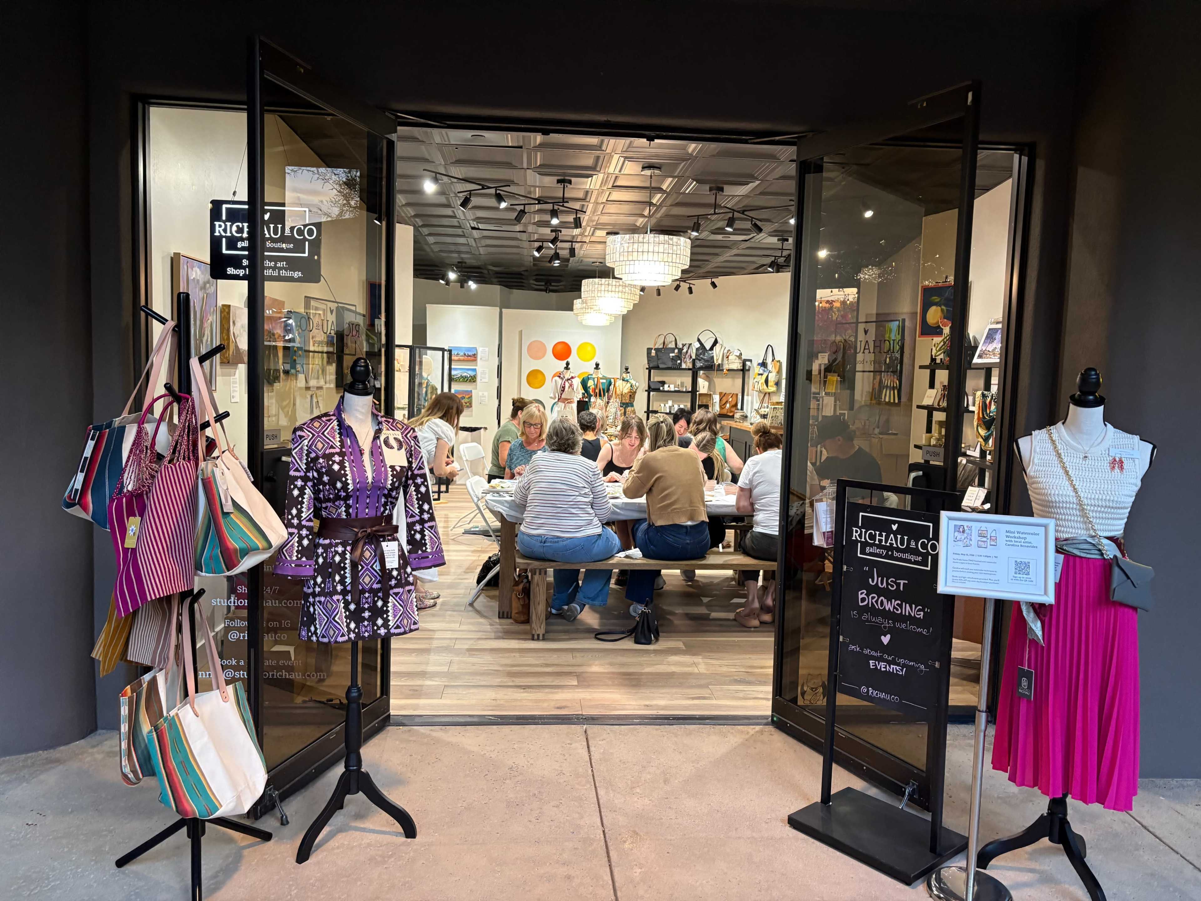A group of people is seated around a long table inside a shop, while various bags and clothing items are displayed at the entrance.