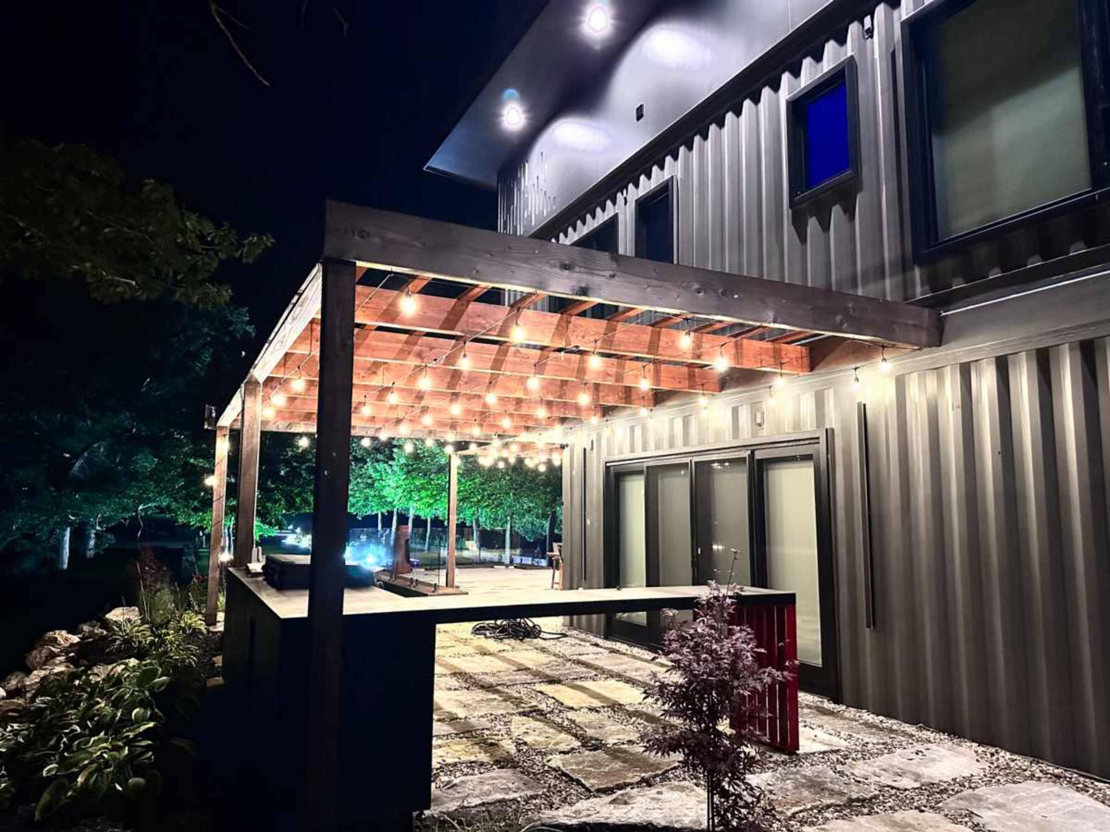 The image shows a modern outdoor patio area with a wooden pergola illuminated by string lights, adjacent to a sleek building.