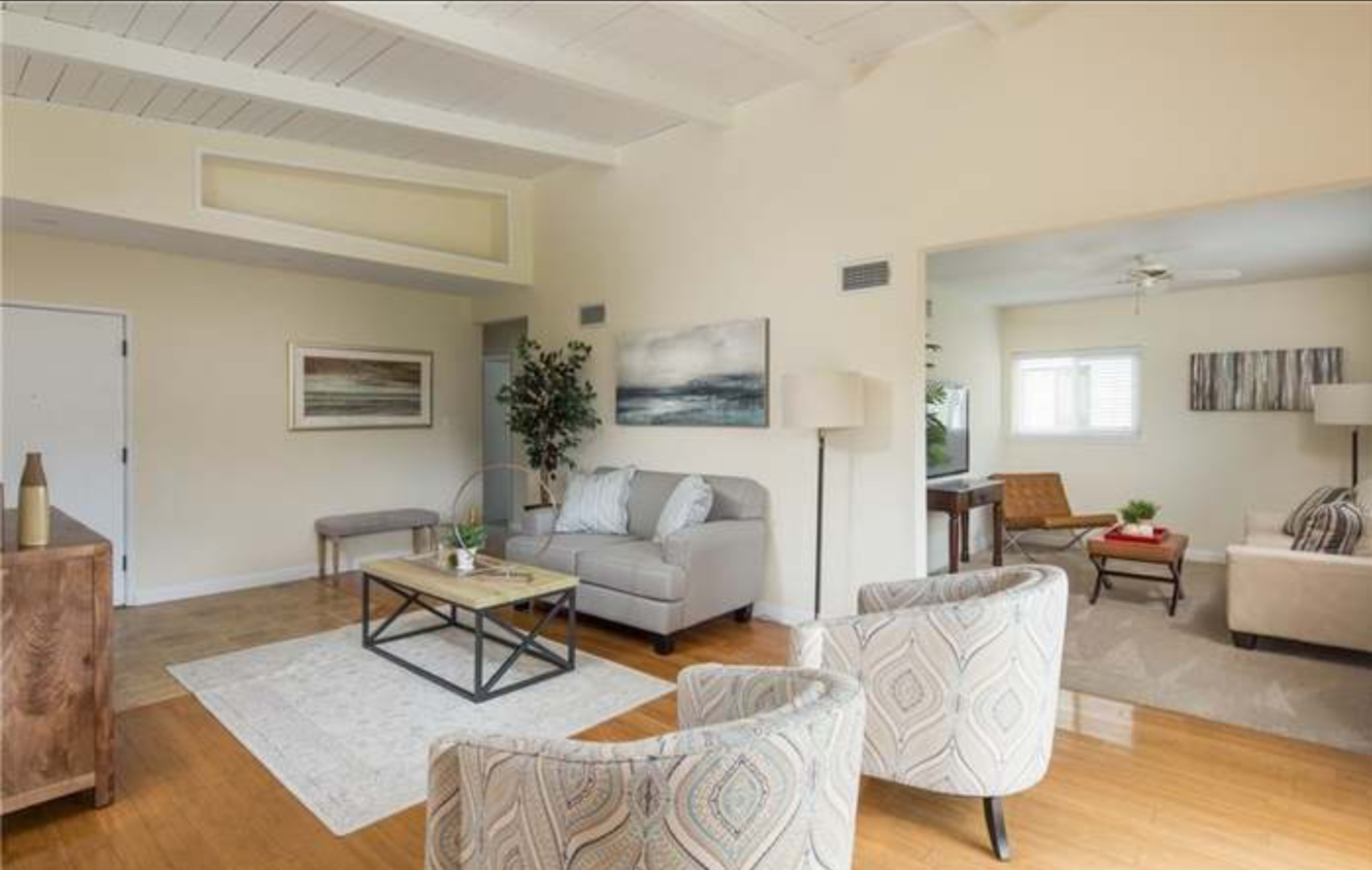 A bright living room with light-colored walls, a gray sofa, two patterned chairs, a coffee table, and a small dining area visible in the background.