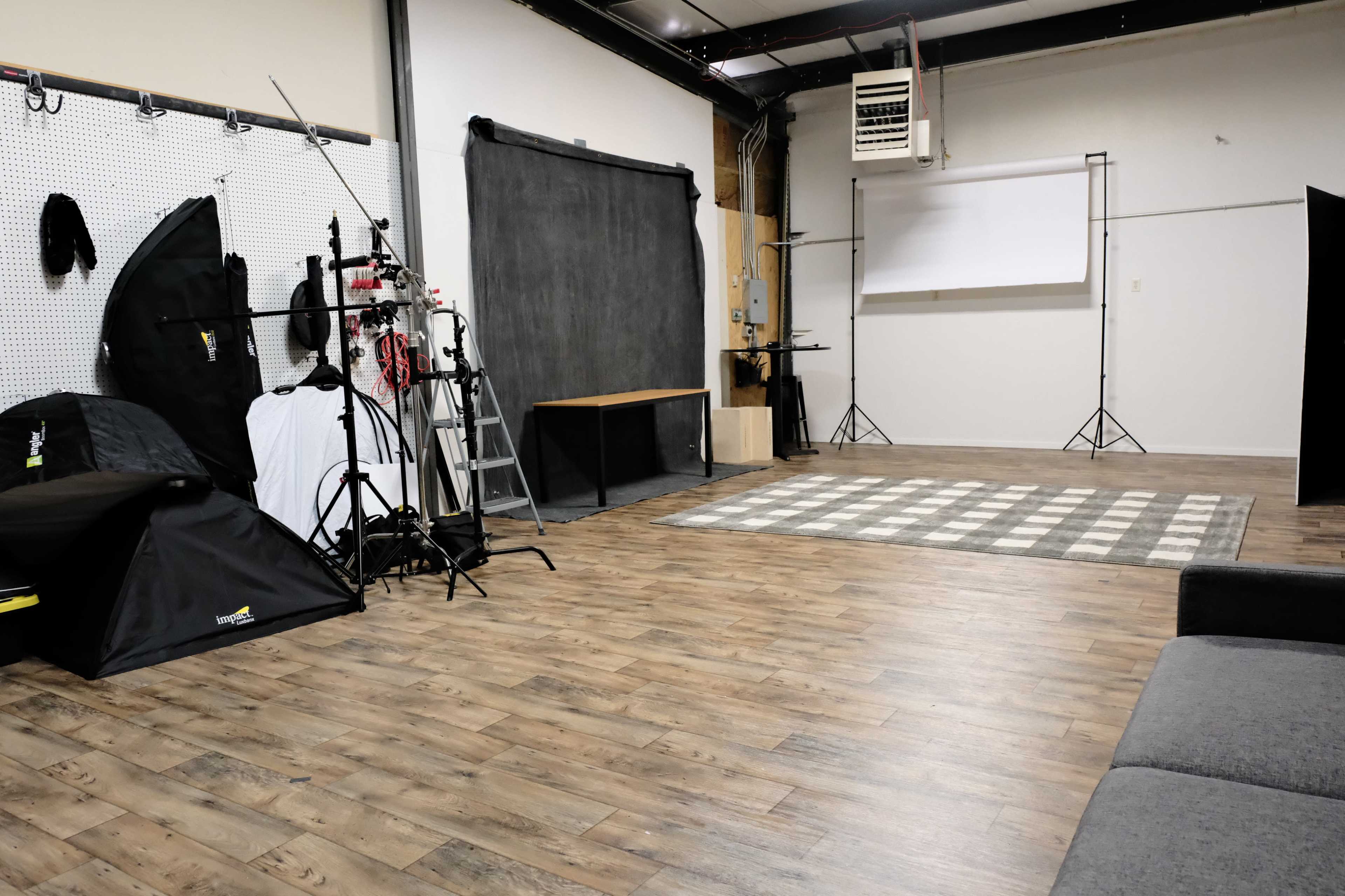A spacious photography studio with wooden flooring, a checkered rug, and various equipment including backdrop stands and lighting setups against the walls.