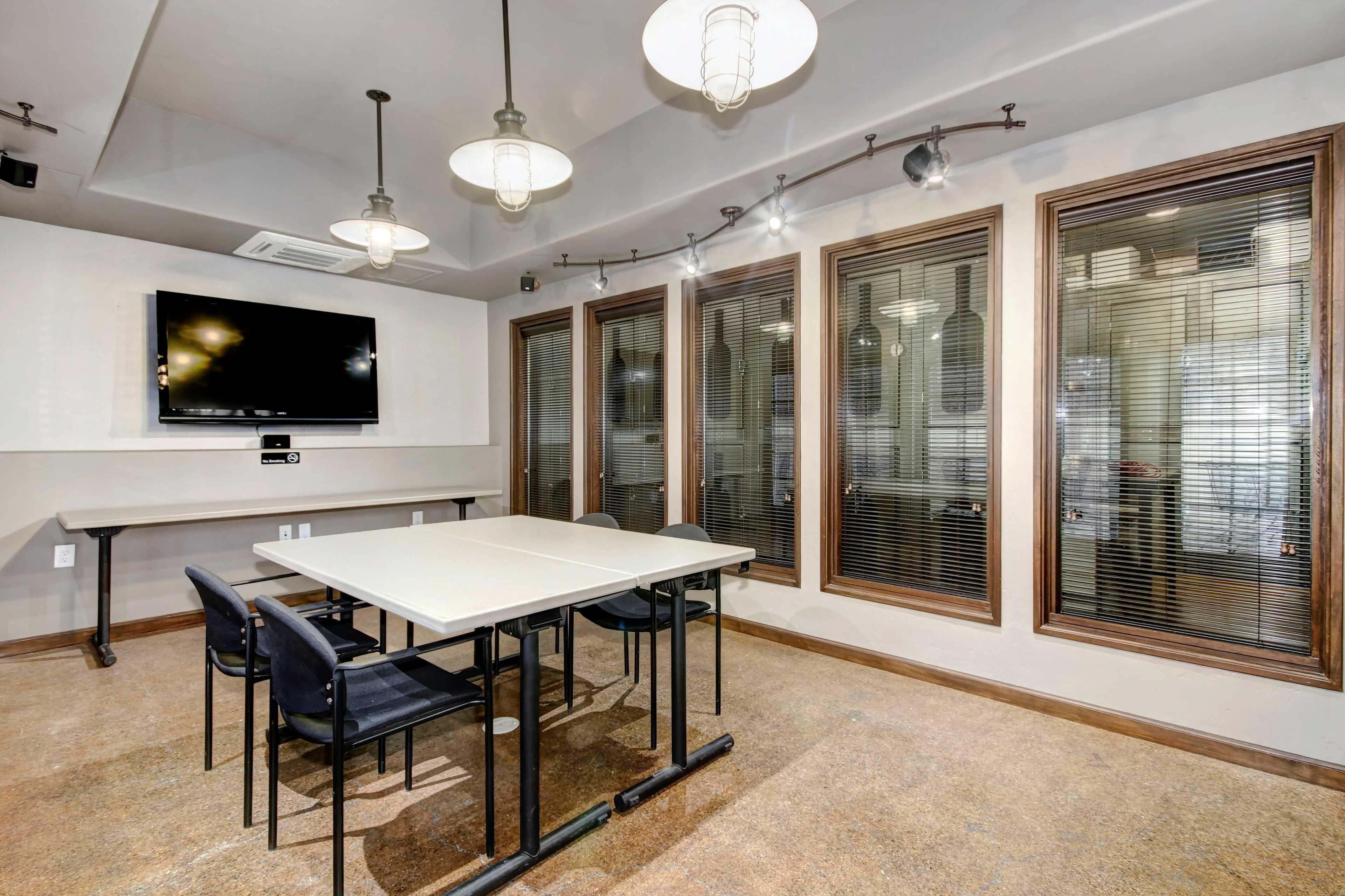 A conference room features a rectangular table surrounded by black chairs, a wall-mounted screen, and large windows with blinds.