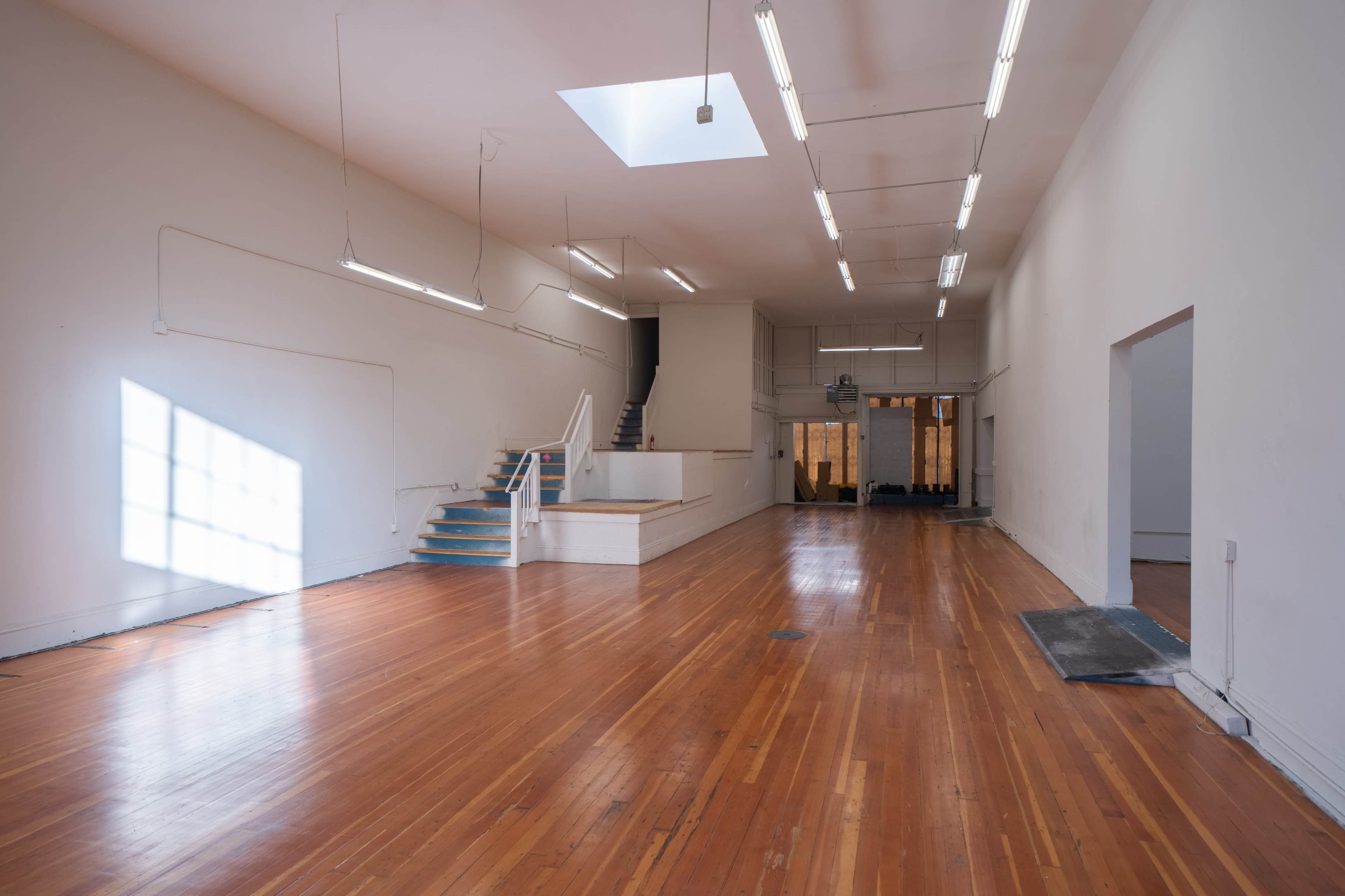 A large, empty room with polished wooden floors, high ceilings, and a skylight, featuring a staircase at the back and walls devoid of furnishings.