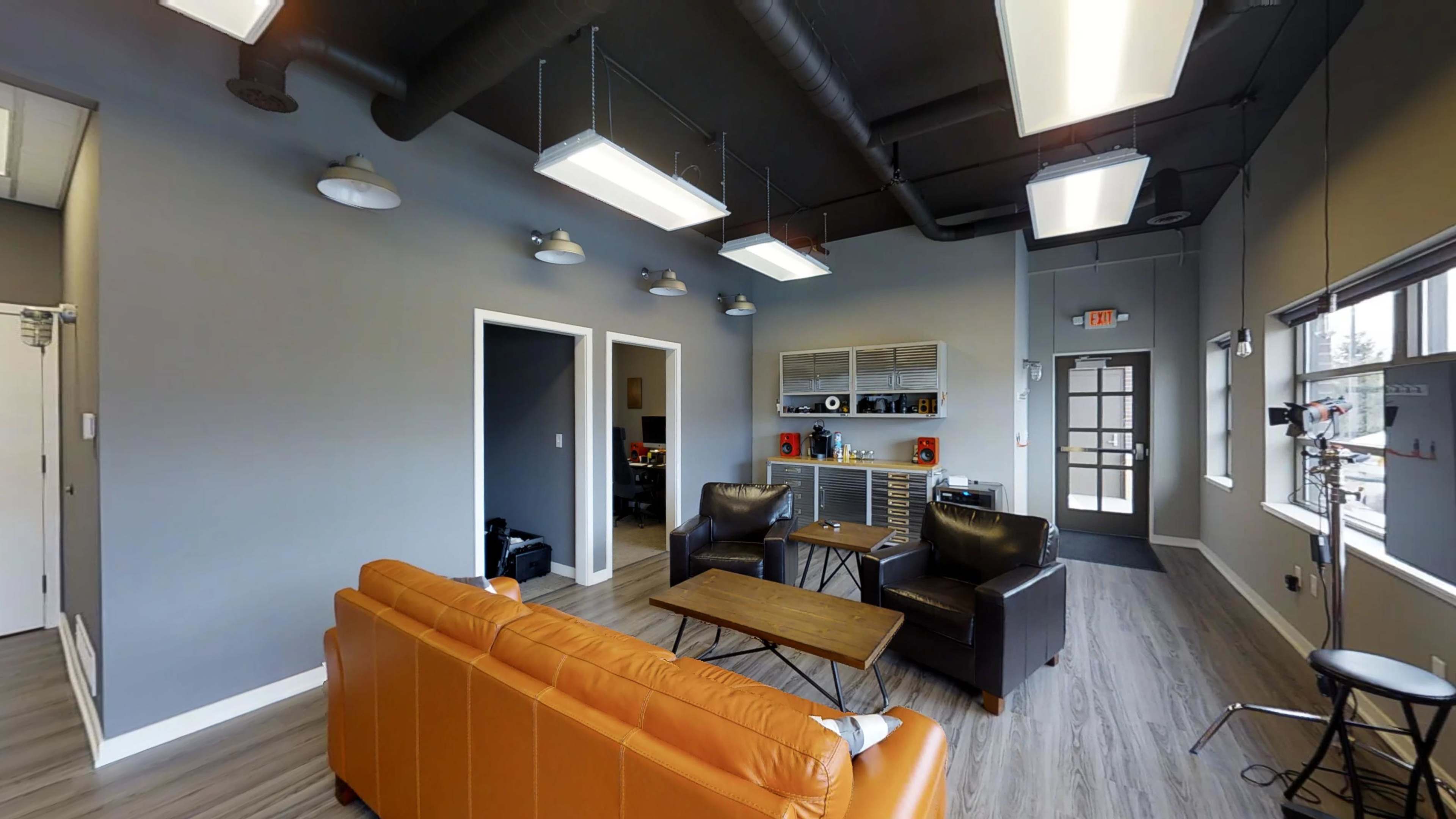 A spacious office lounge area with an orange couch, two black leather chairs, a wooden coffee table, and overhead lighting.