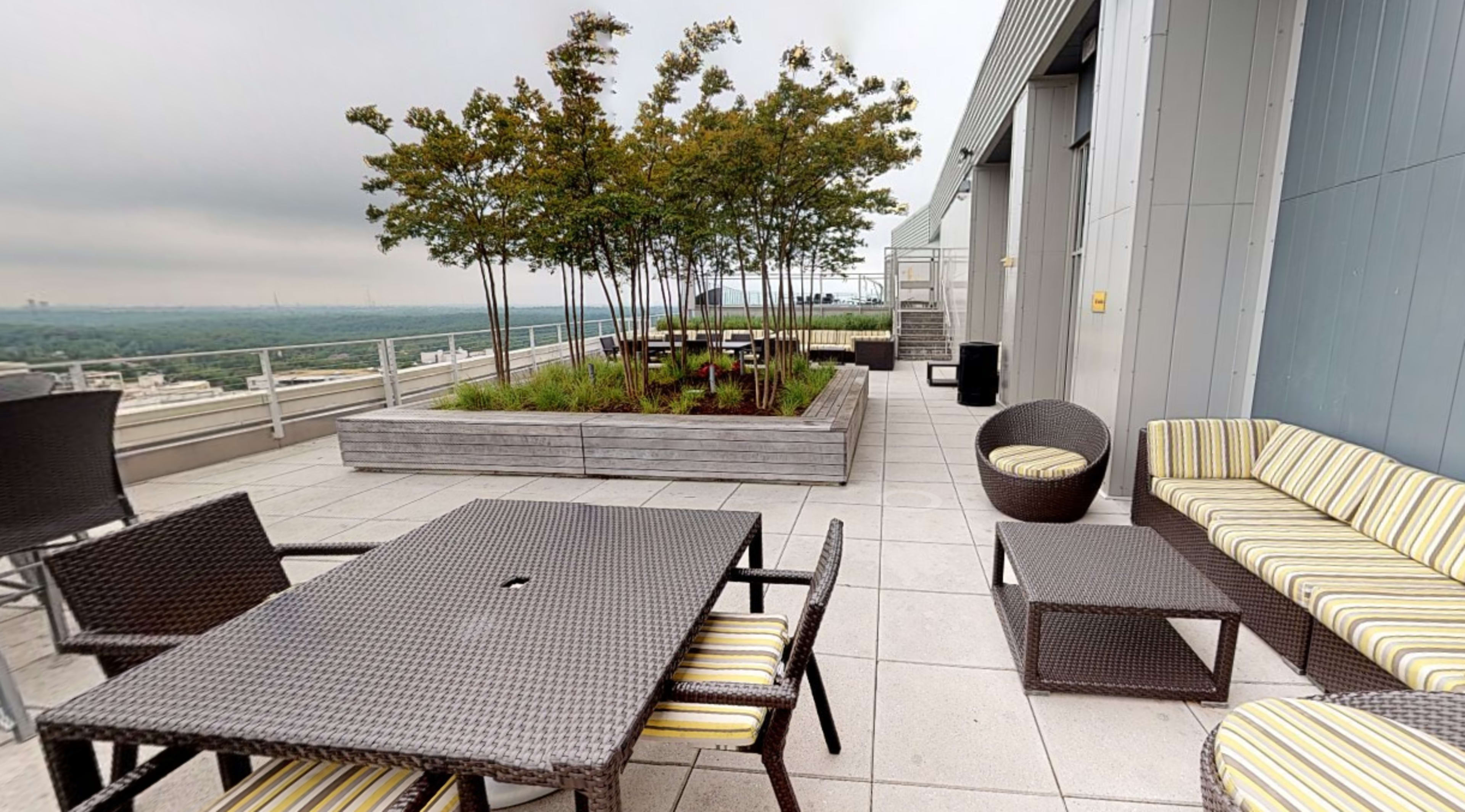 A rooftop terrace featuring outdoor furniture, including tables and seating, surrounded by planters with trees.