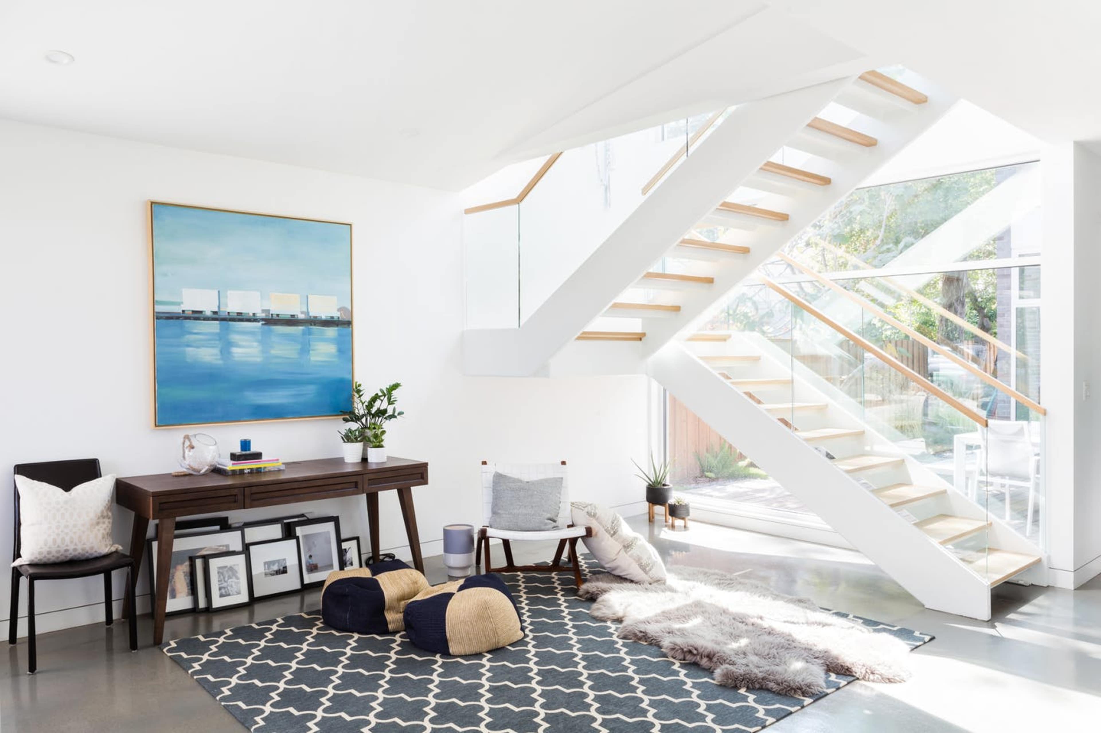 A modern interior featuring a staircase, a desk with a plant, and a seating area with poufs on a patterned rug.