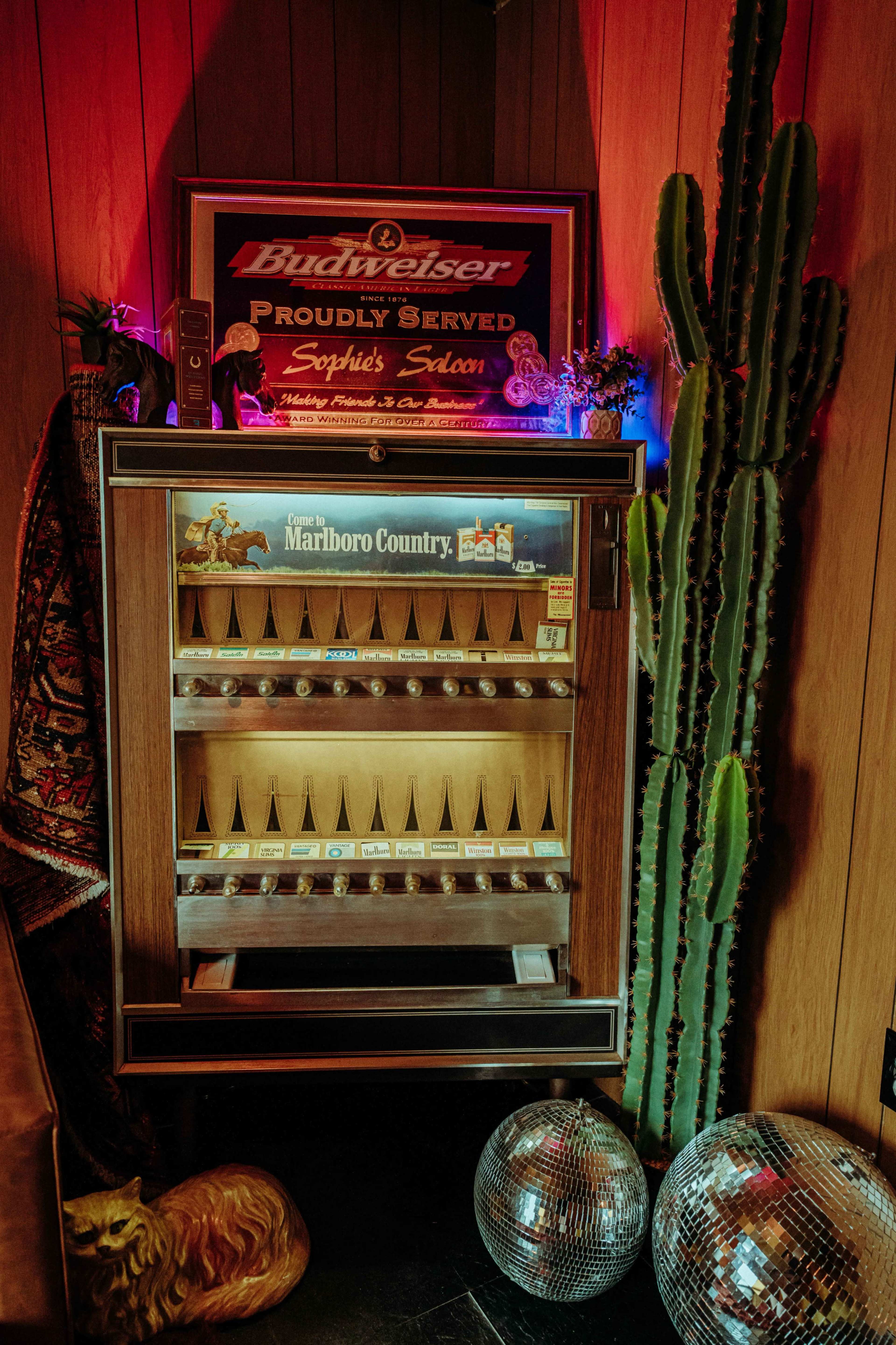 A vintage vending machine filled with boxed goods is displayed alongside a cactus and decorated with colorful lights and disco balls.