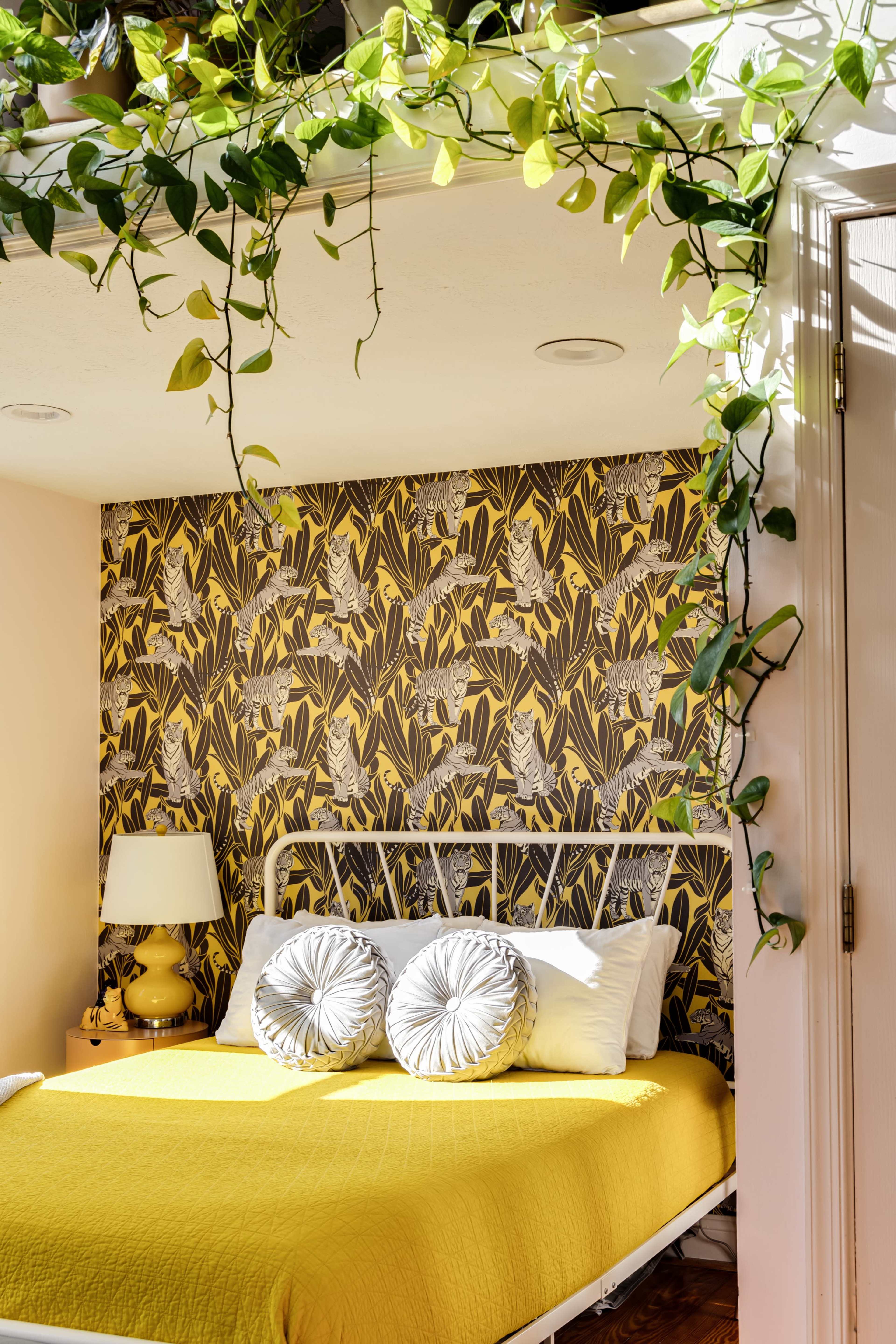 A bedroom features a yellow bedspread, decorative pillows, and wallpaper with a botanical pattern, complemented by trailing green plants.