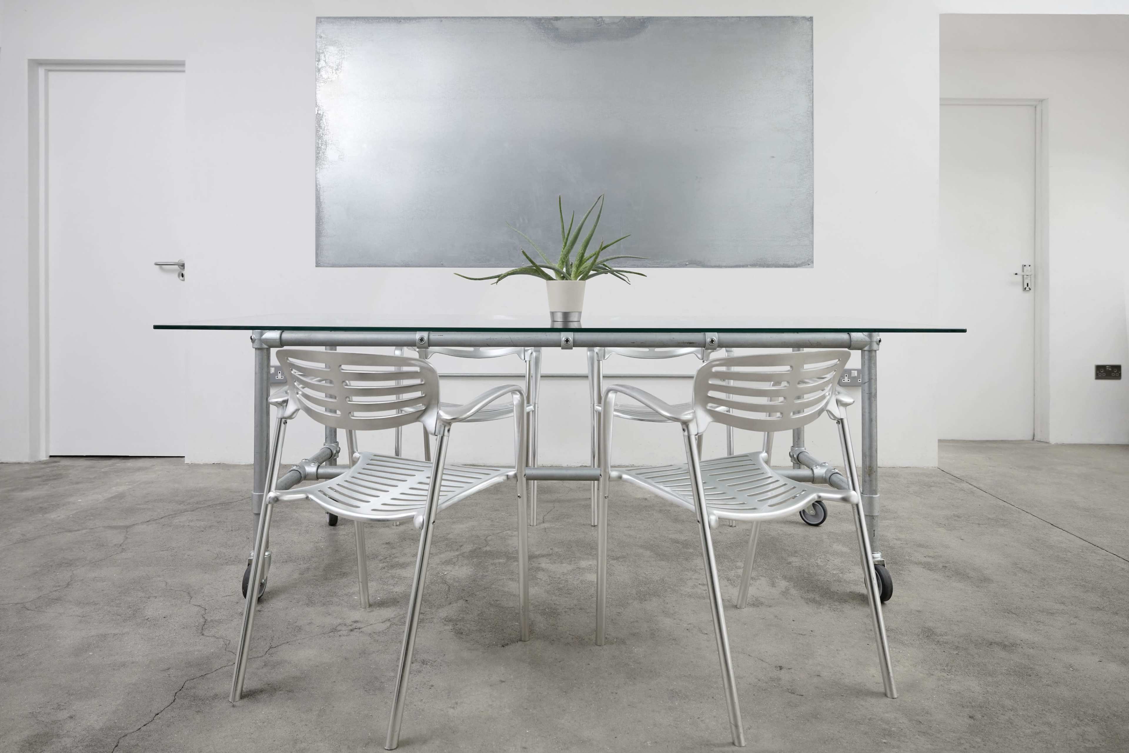 A glass-top table surrounded by four white metal chairs is placed on a concrete floor in a minimalist room with gray walls.