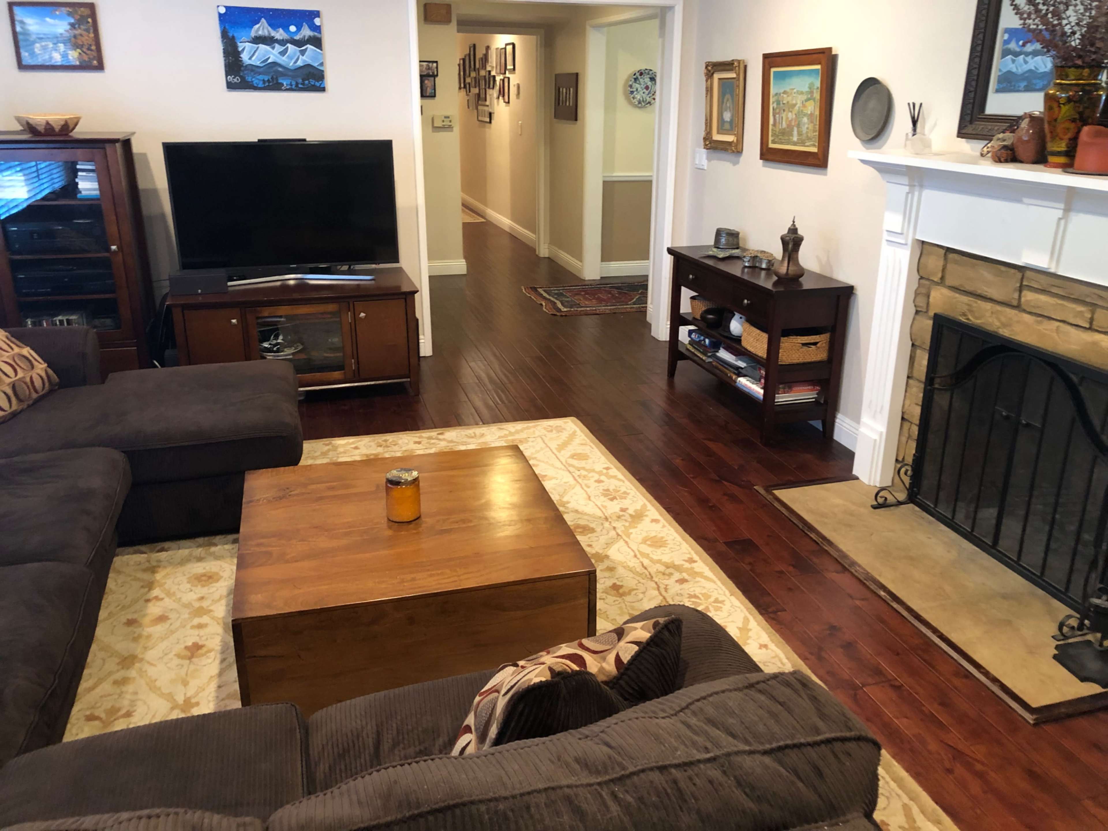 The image shows a cozy living room with a dark sectional sofa, a wooden coffee table, a television cabinet, and a fireplace with artwork on the walls.