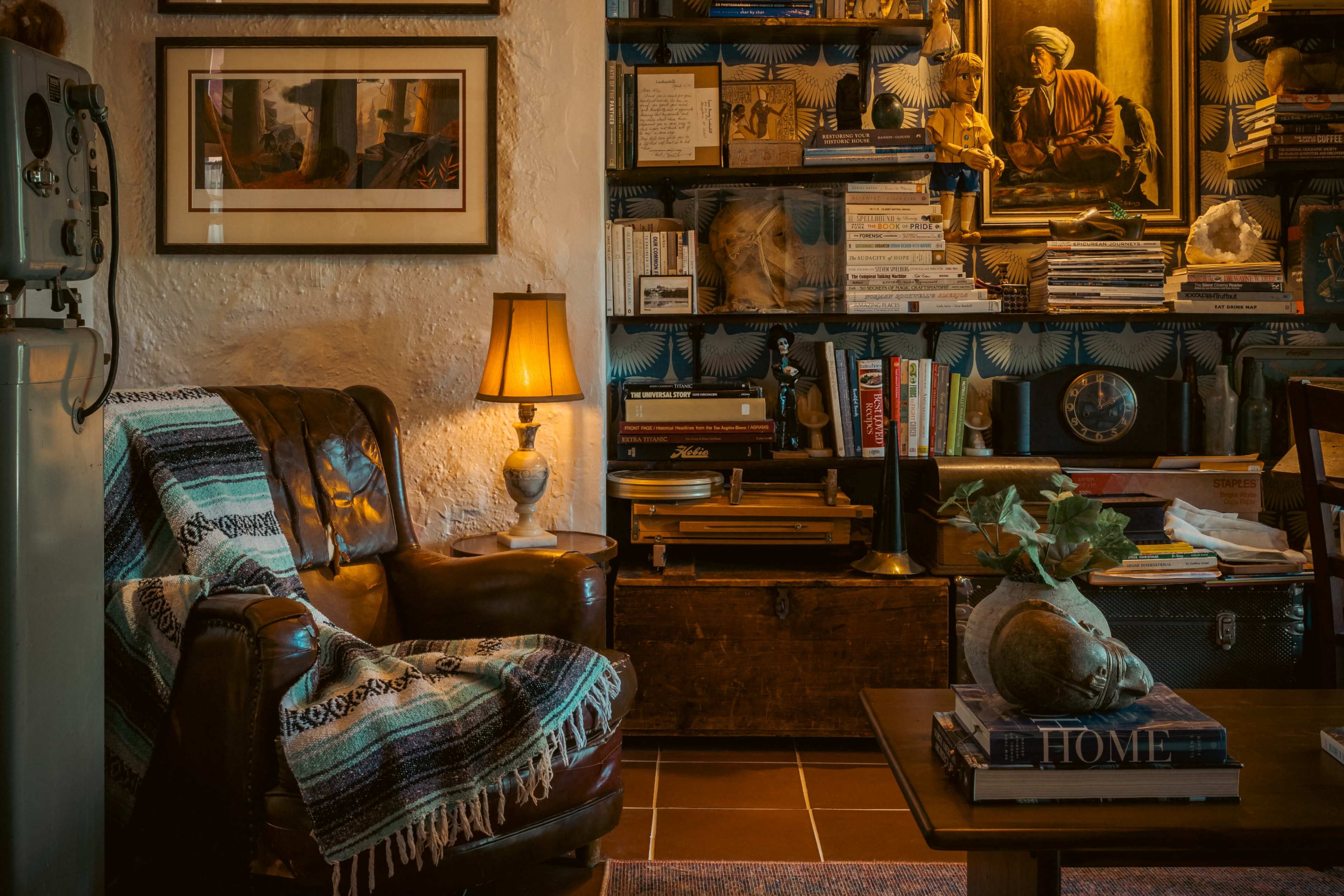 The image shows a cozy living room featuring a leather armchair adorned with a patterned blanket, a side table with a lamp, and a bookshelf filled with books and decorative items.