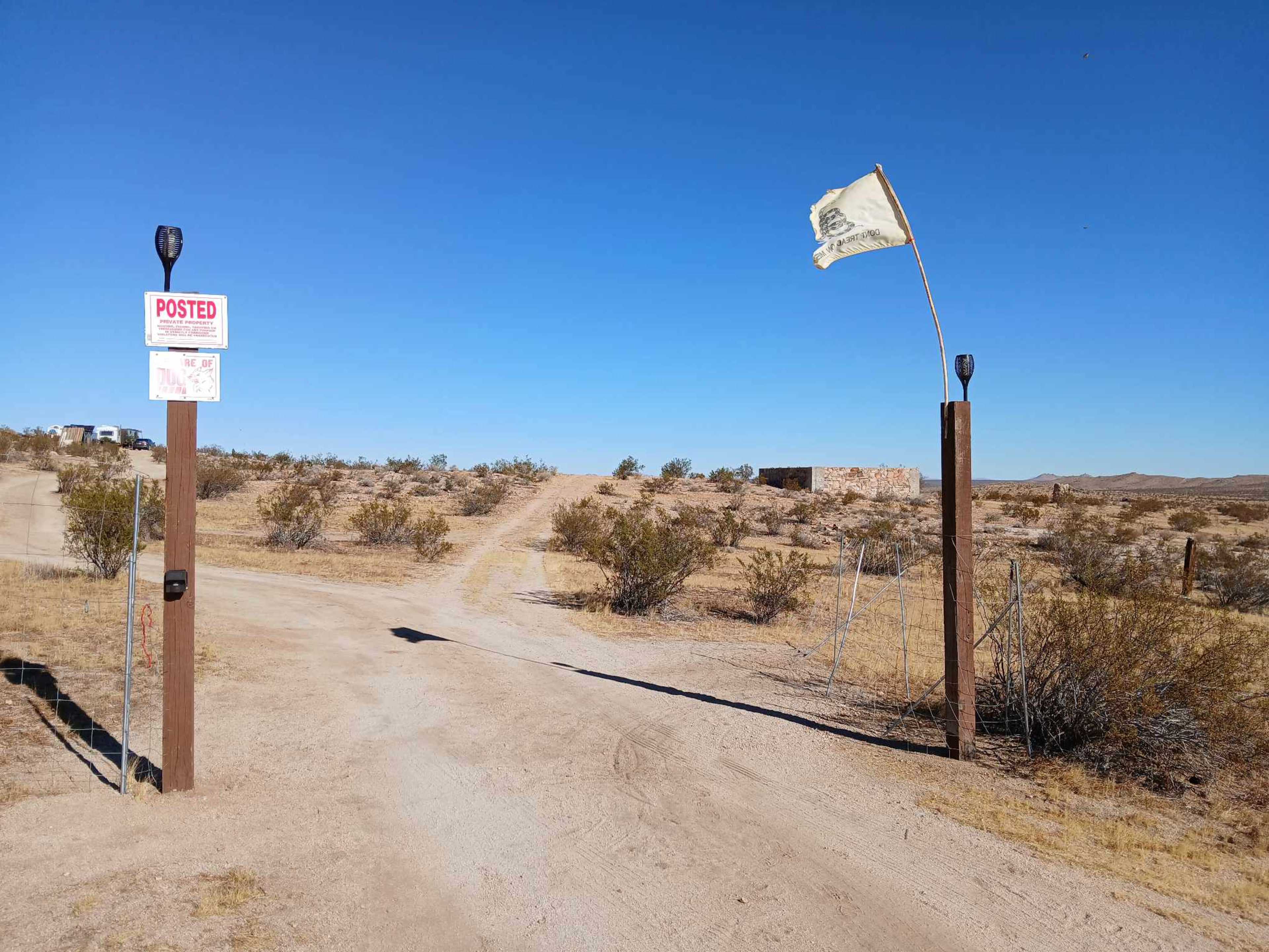 A dirt road leads into a sparsely vegetated area, marked by a "Posted" sign on a wooden post, with a flag waving nearby.