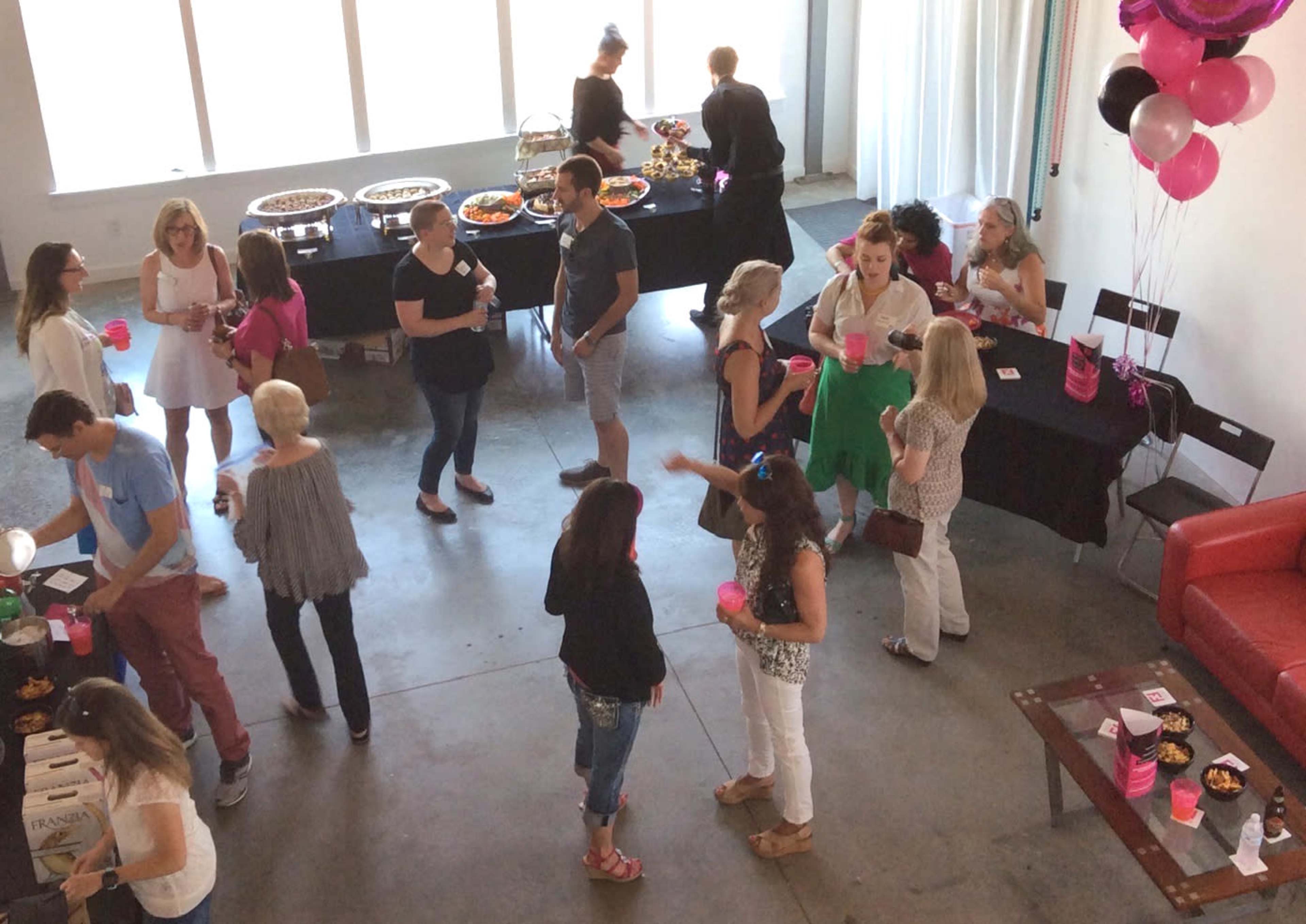 A group of people socializes at a festive event in a well-lit venue, with tables arranged for food and drinks.