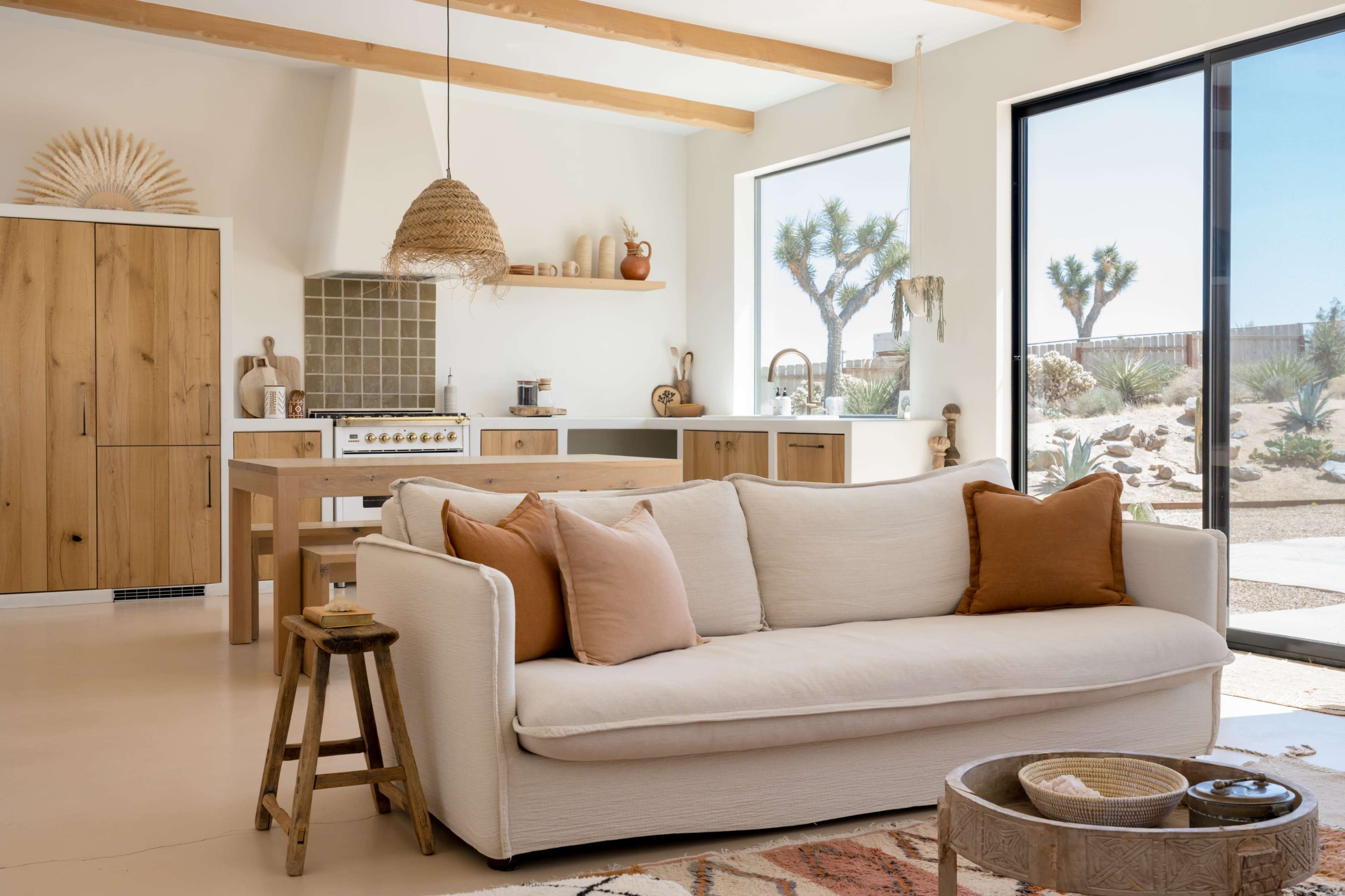 A light-filled living area features a cream sofa with decorative pillows, a wooden dining table, and large glass doors opening to a desert landscape.