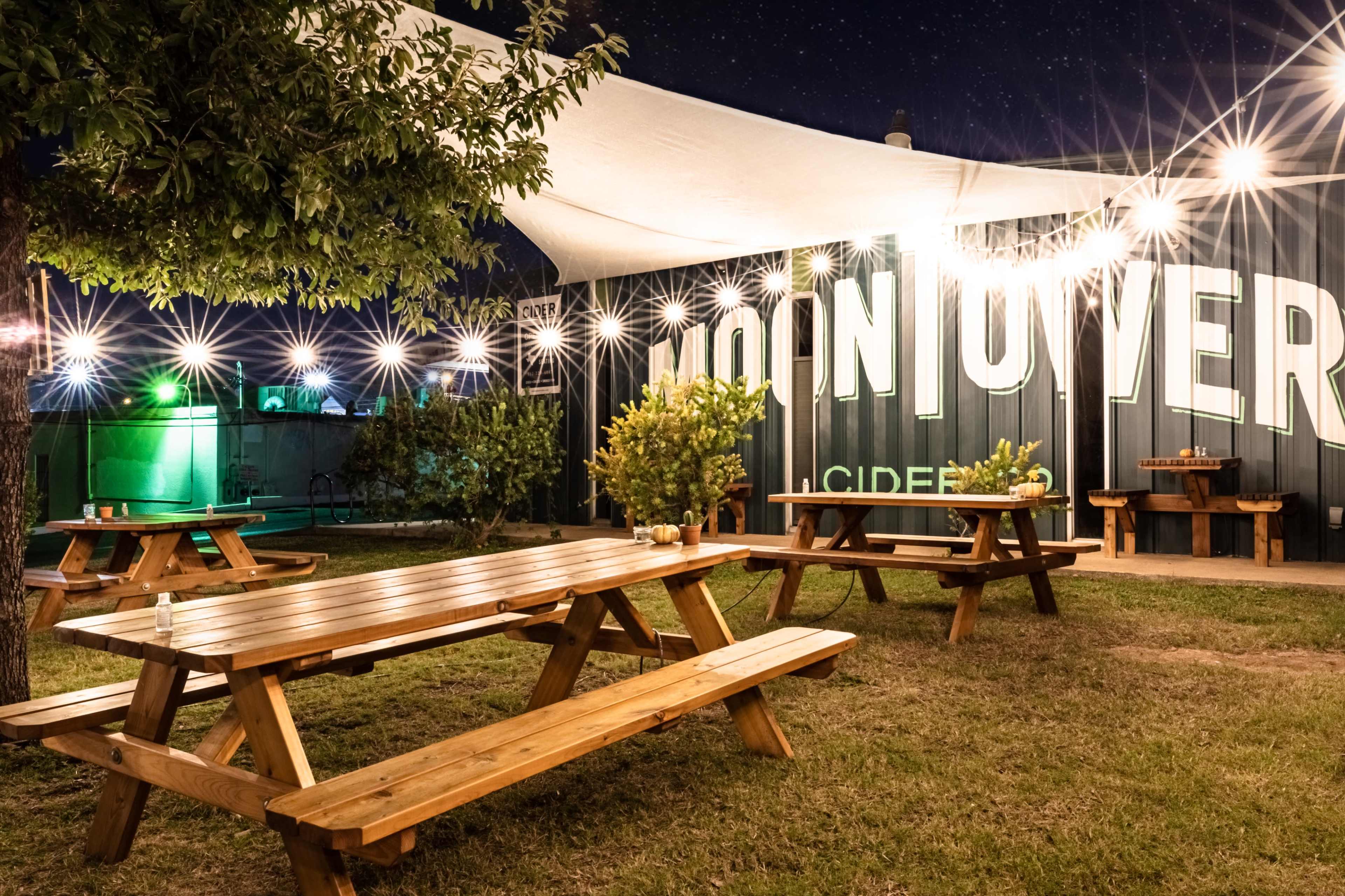 A wooden picnic area under a canopy with string lights illuminating the space, surrounded by greenery and a large sign that reads "MOON OVER."
