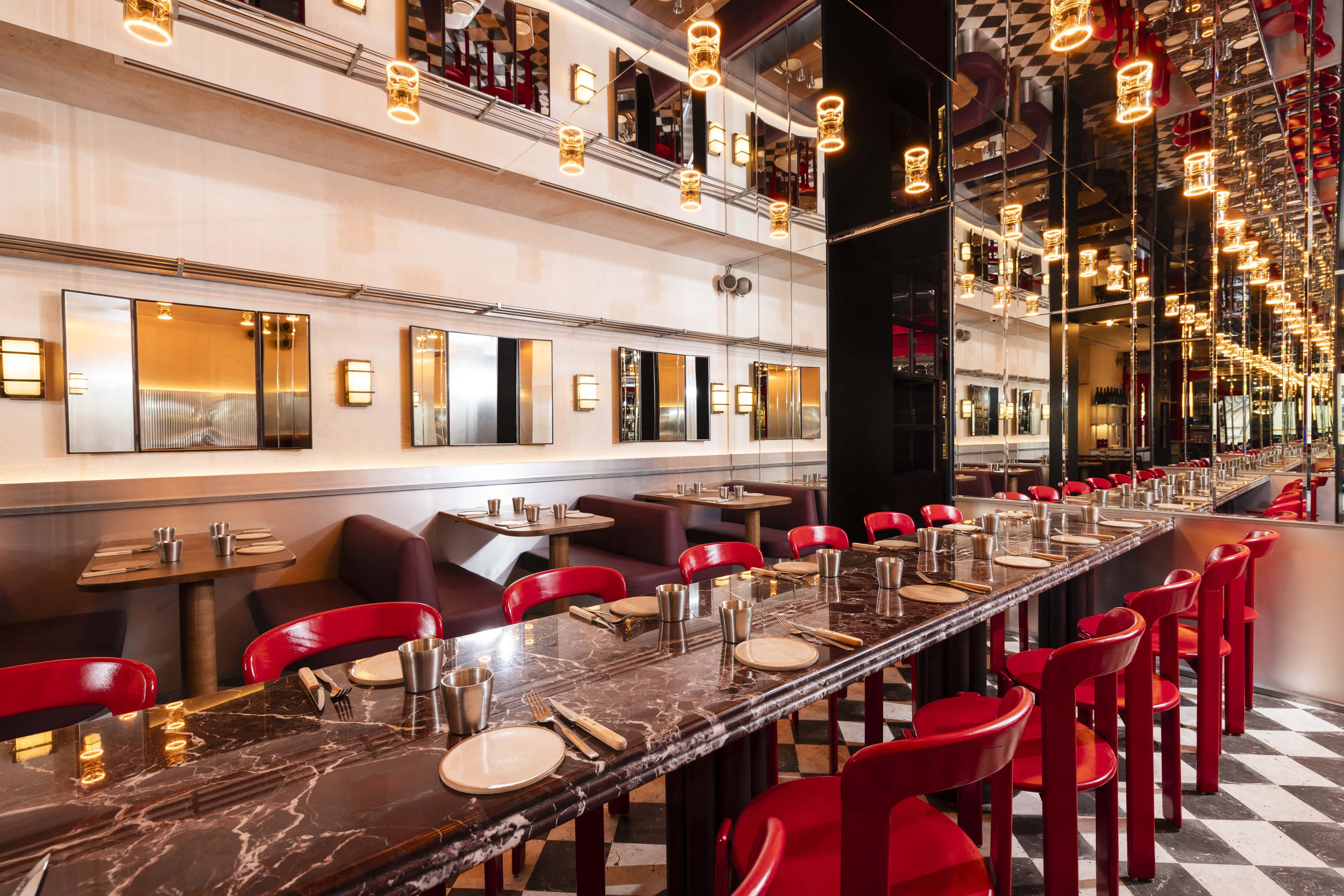 A modern restaurant interior features a long marble table lined with red chairs, illuminated by pendant lights, with mirrored walls reflecting the vibrant decor.