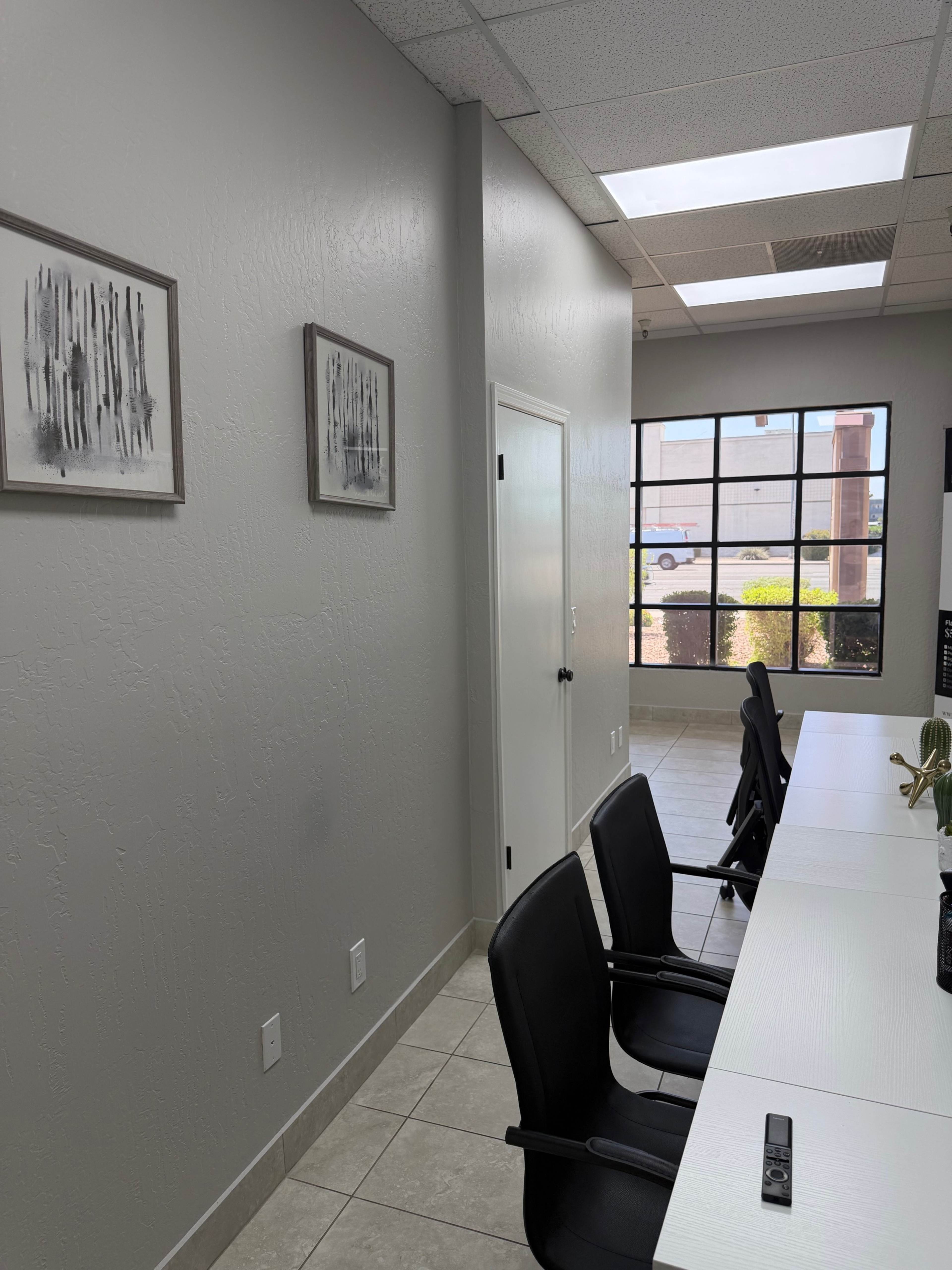 The image shows a modern office interior with a long white desk facing several black chairs, and light gray walls adorned with framed artwork.