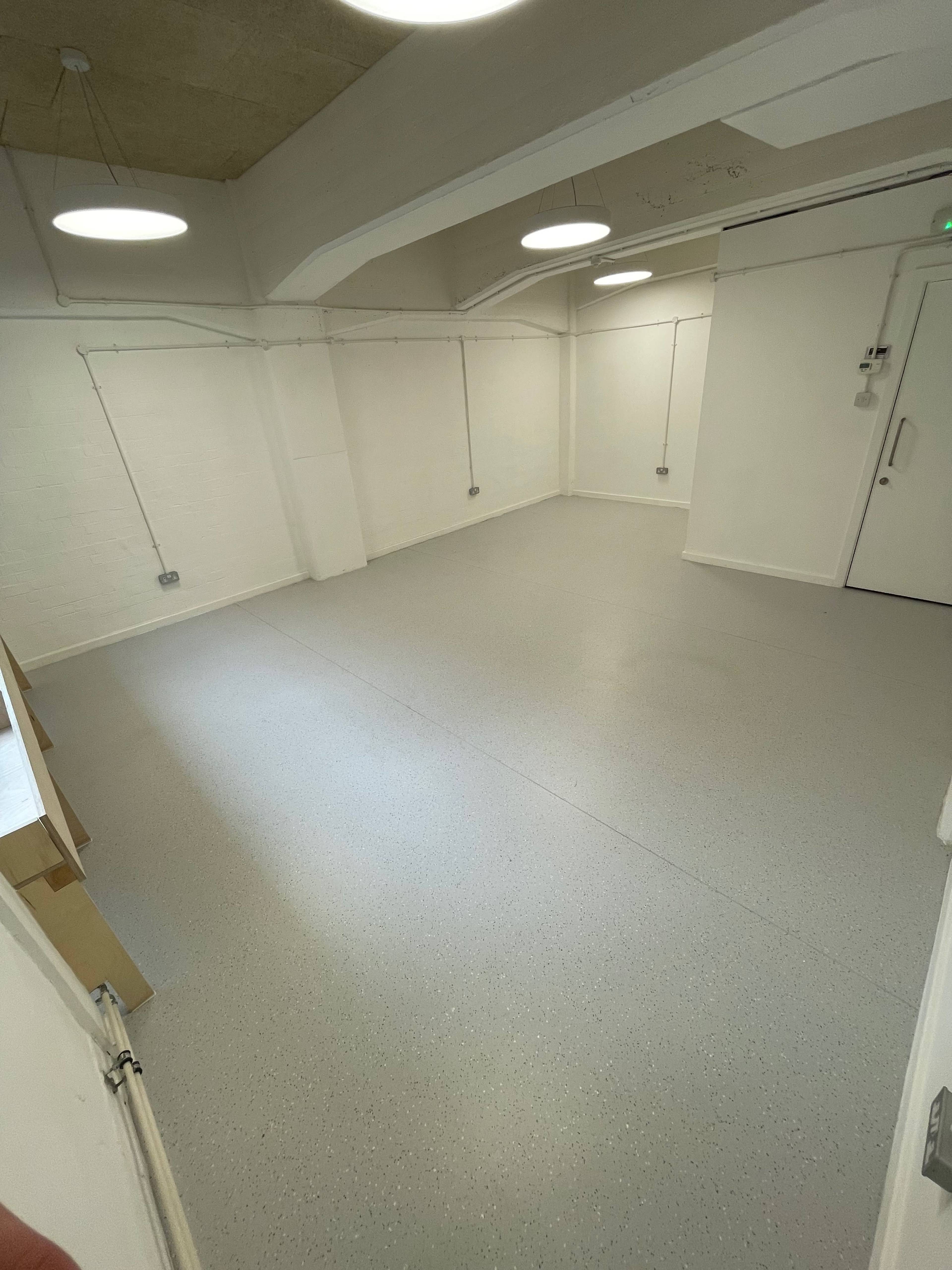 An empty room with a gray floor and white walls, featuring overhead circular lights and a single door.
