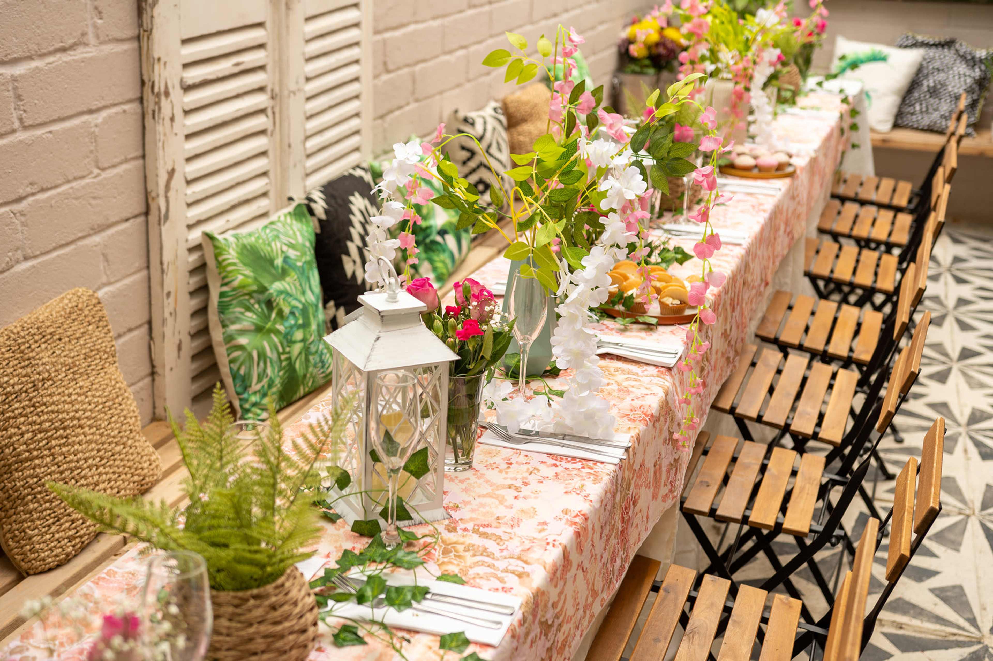 A long dining table is set with floral arrangements and decorative items, flanked by wooden chairs in a brightly lit room.