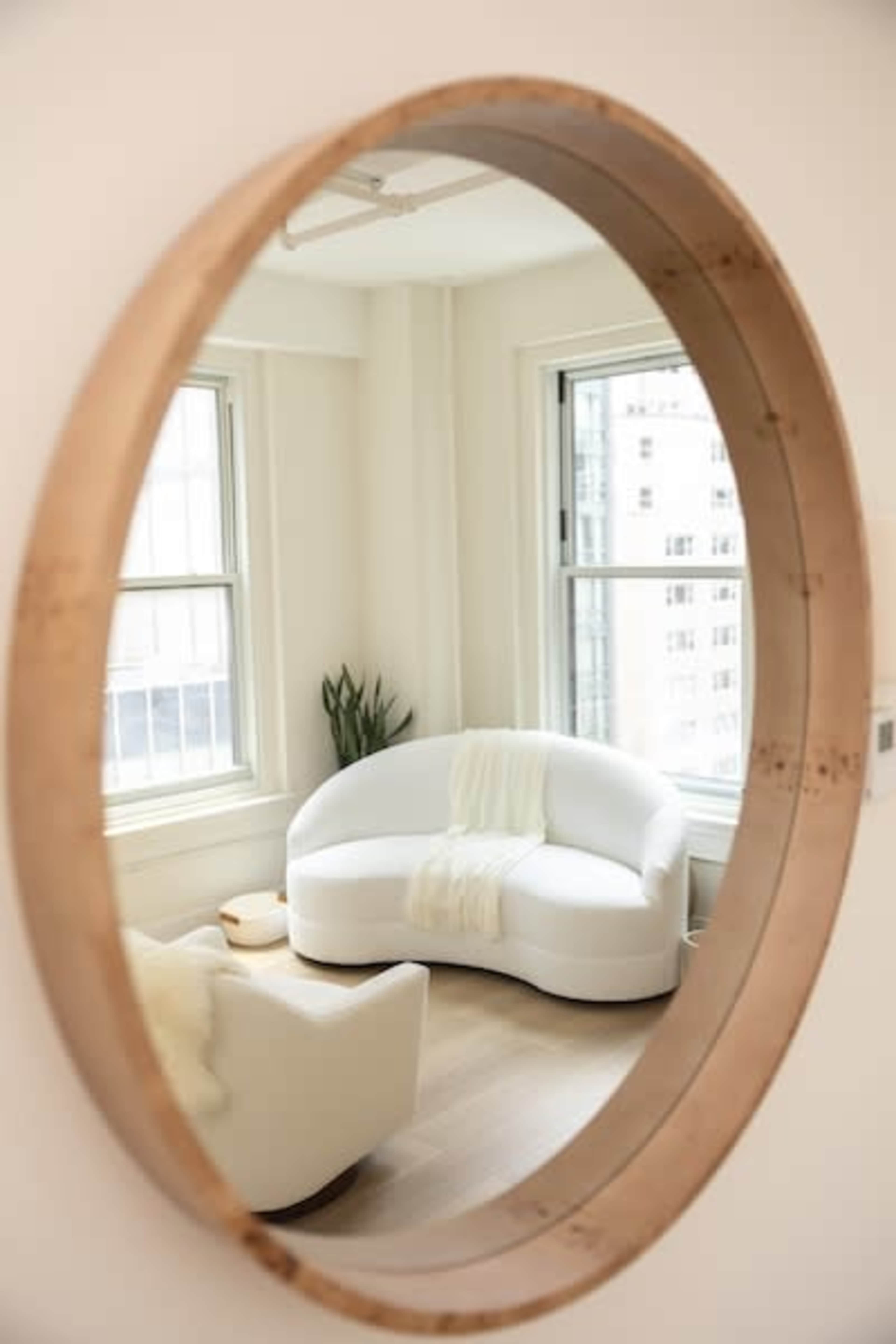 A round wooden-framed mirror reflects a white, curved sofa and a houseplant in a well-lit room with large windows.