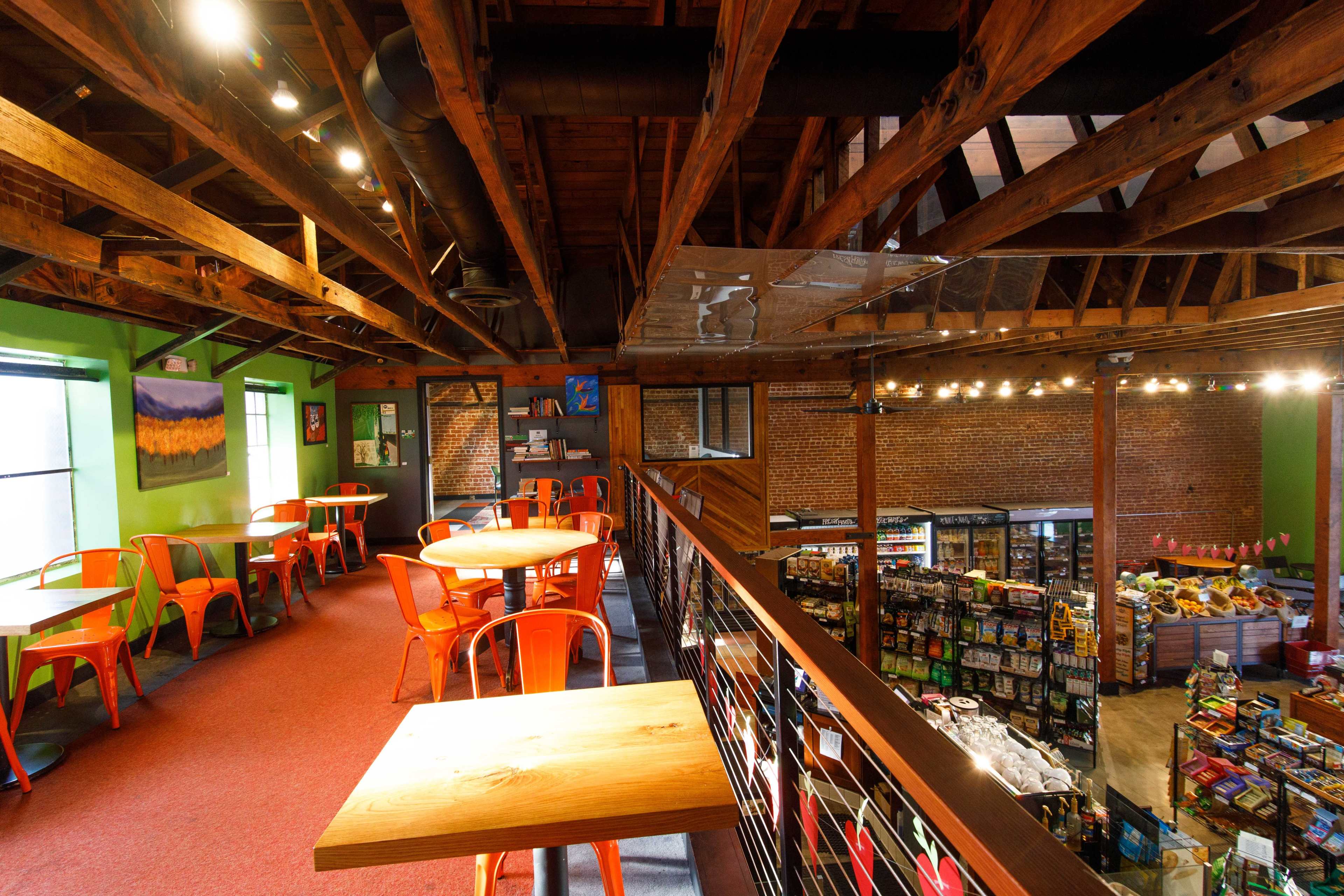 A two-level café and grocery store with wooden beams, orange chairs, and shelves stocked with various food items on the lower level.