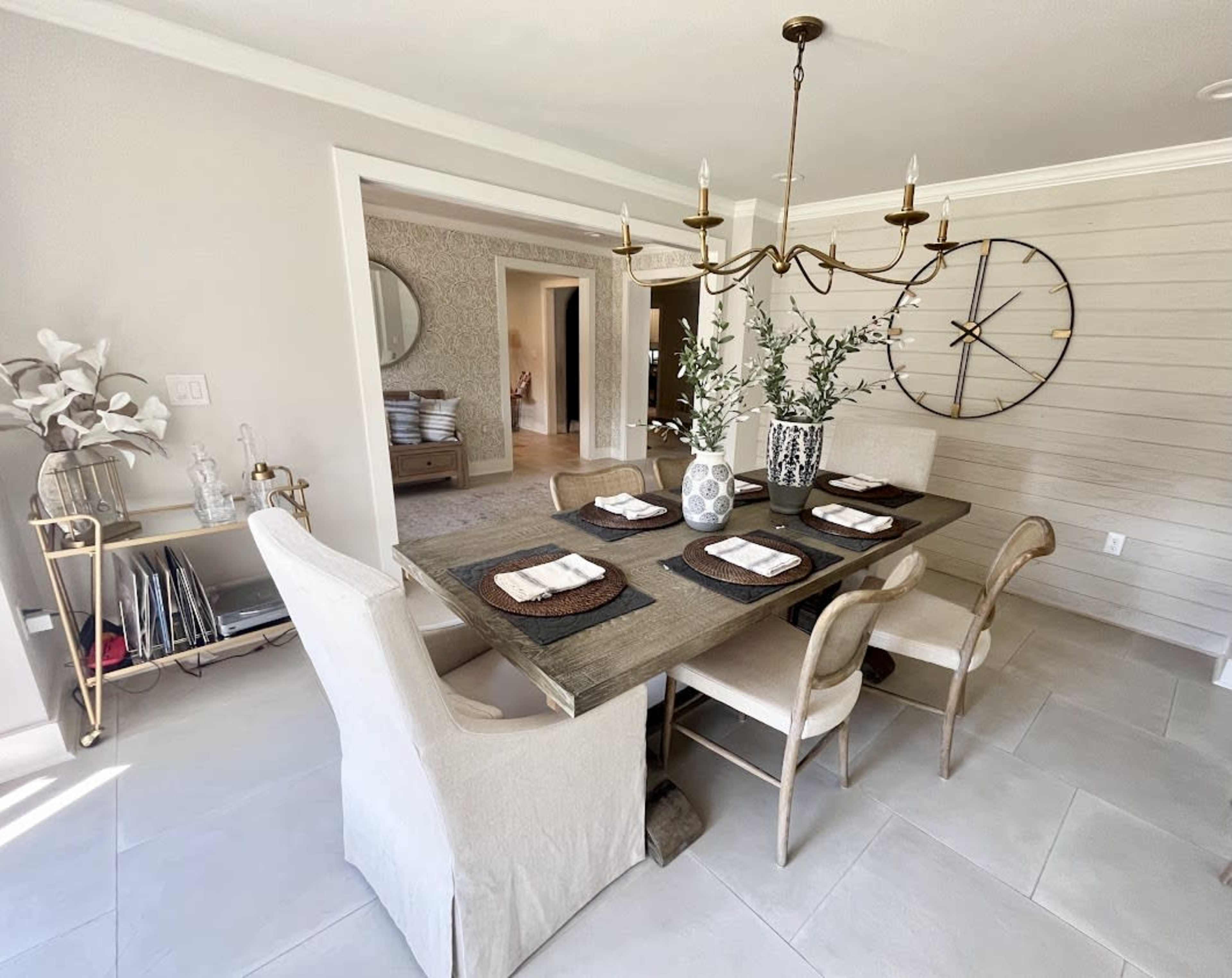 A dining area features a wooden table set with place mats and surrounded by light-colored chairs, with a chandelier above and accessories on a nearby cart.