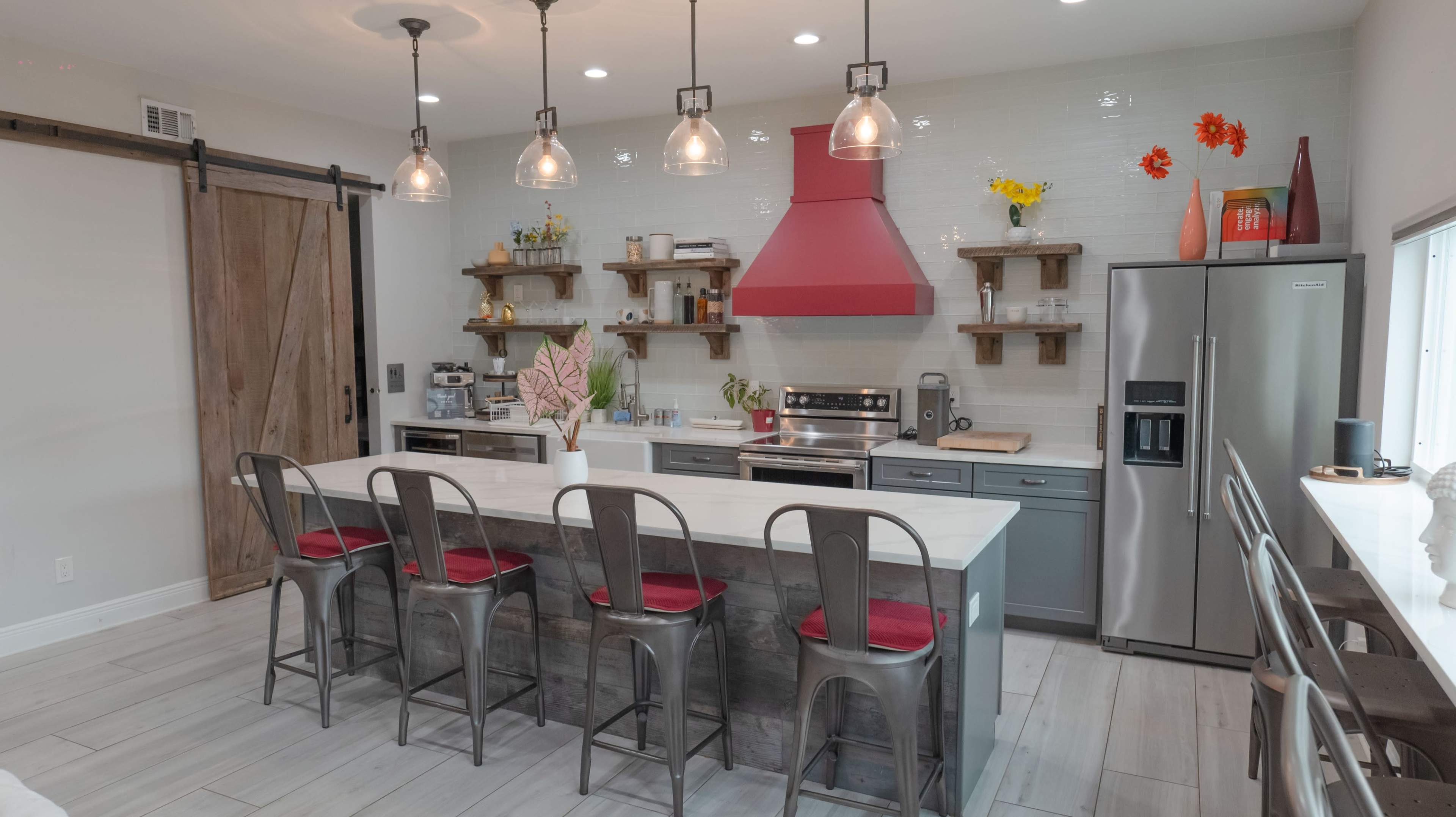 The kitchen features a central island with barstool seating, stainless steel appliances, red accents, and wooden shelves displaying various kitchen items.