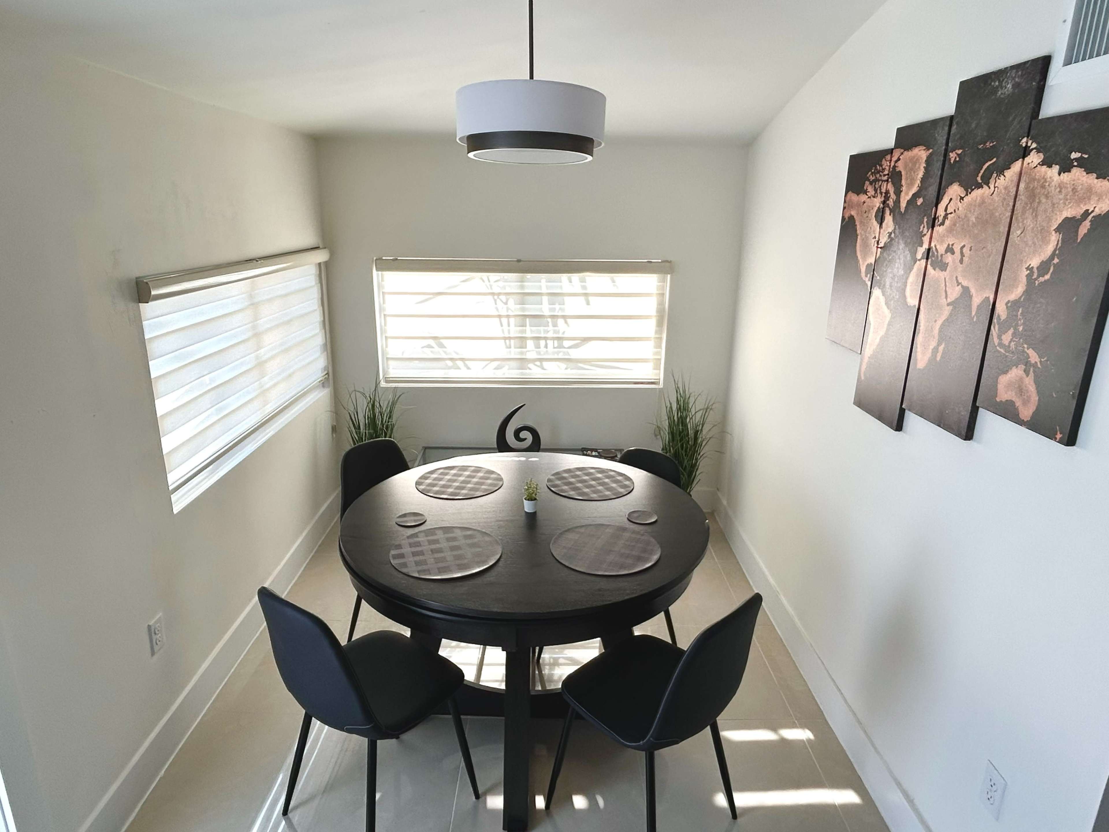 The image shows a round dining table set for four chairs in a well-lit room with two window shades and a world map art piece on the wall.
