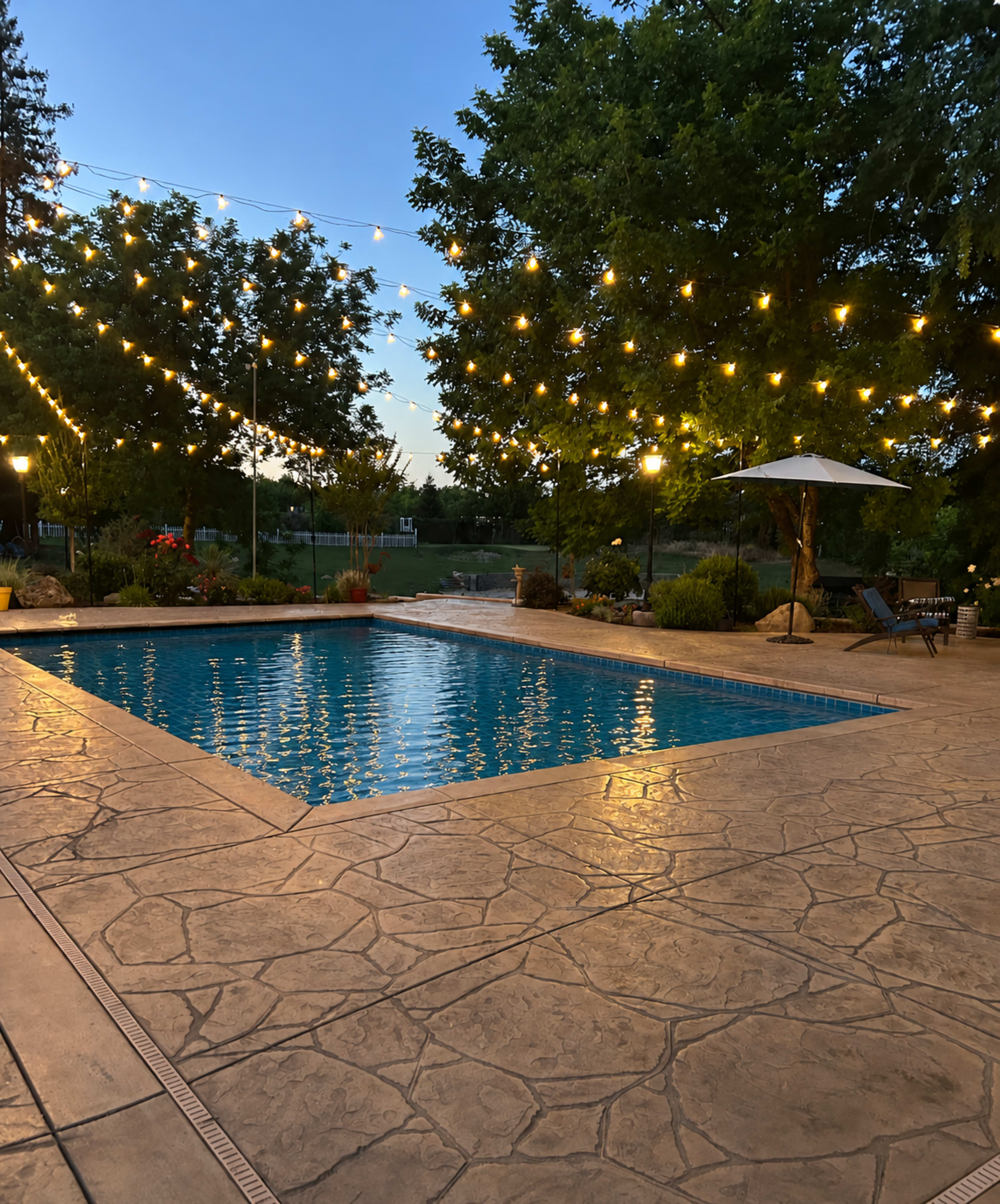 A well-lit swimming pool is surrounded by decorative string lights and landscaped trees in a backyard setting during twilight.